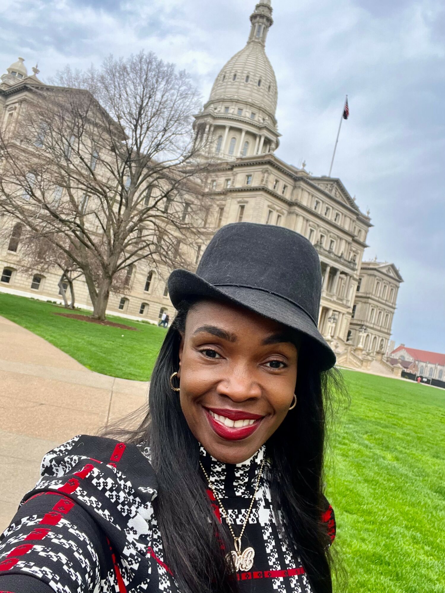 Smiling woman wearing a black hat and patterned top outdoors with a historic building and trees in the background.