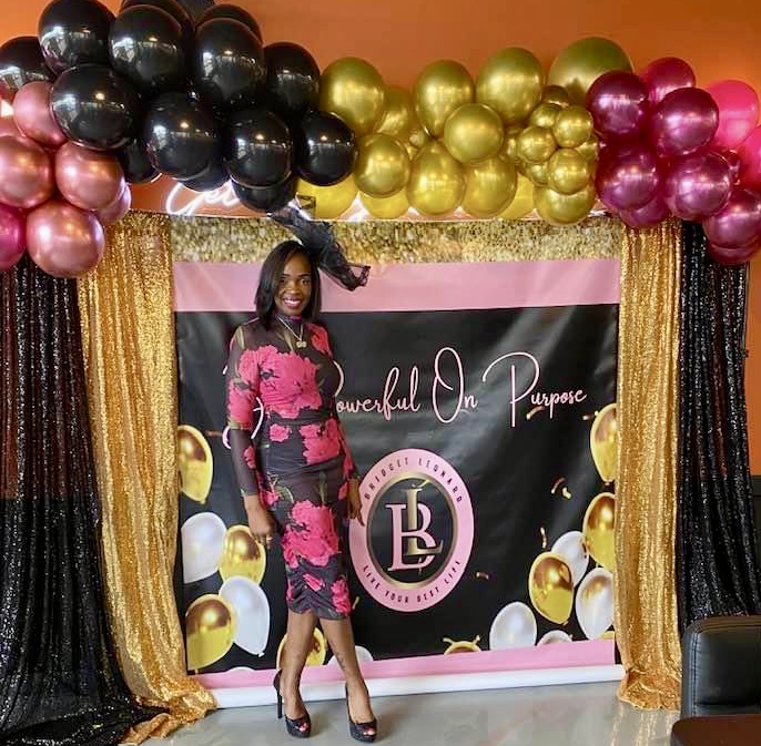 Woman in pink floral dress standing in front of decorated backdrop with balloons and gold curtains.