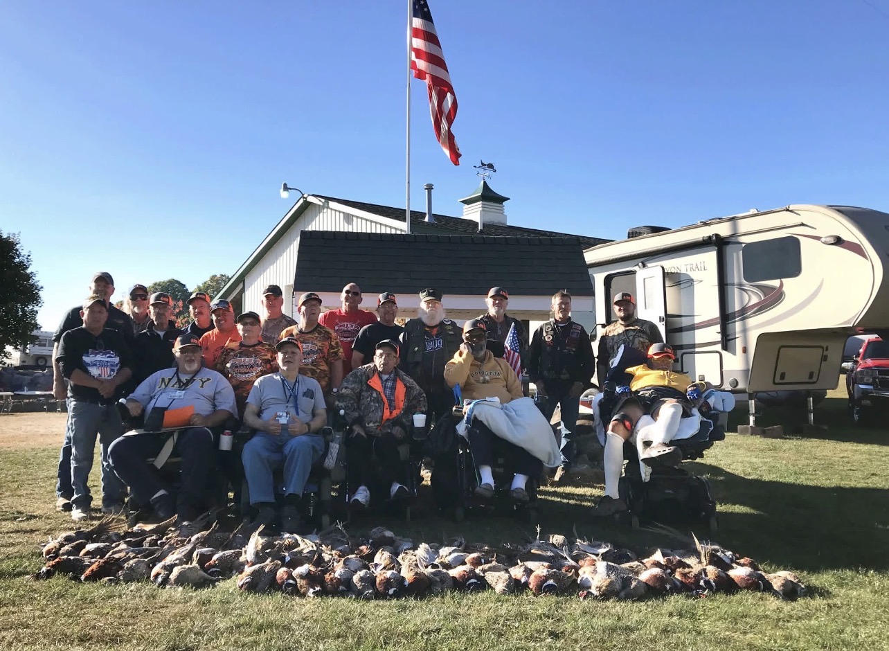 Group of people outdoors with a building, American flag, and RV in background, posing with animals on the ground.
