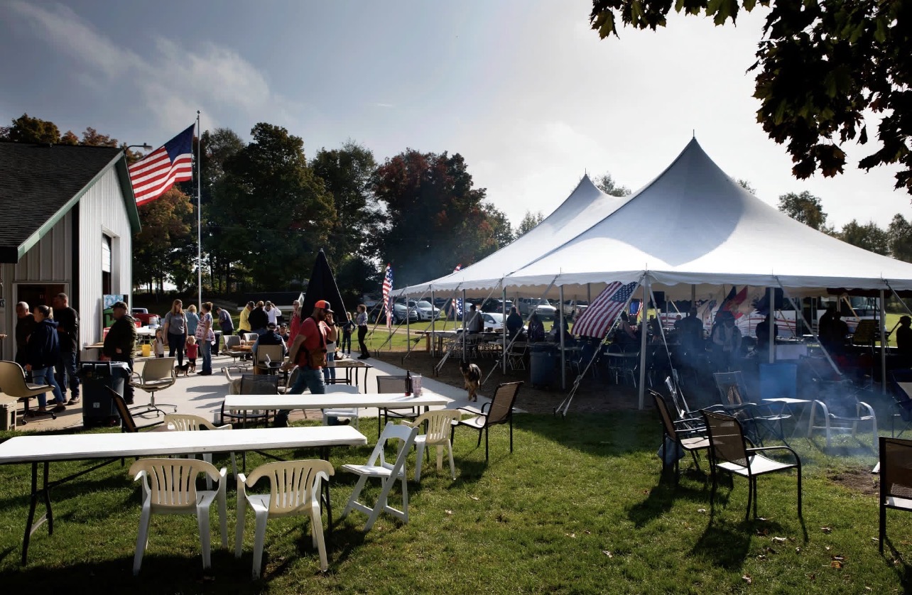 Outdoor gathering with tents, tables, chairs, and people, with trees and a building in the background under a partly cloudy sky.