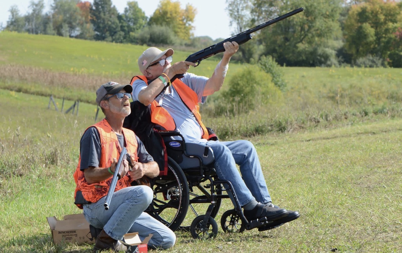 Person in a wheelchair holding a fishing rod, with another person kneeling beside them outdoors in a grassy field.