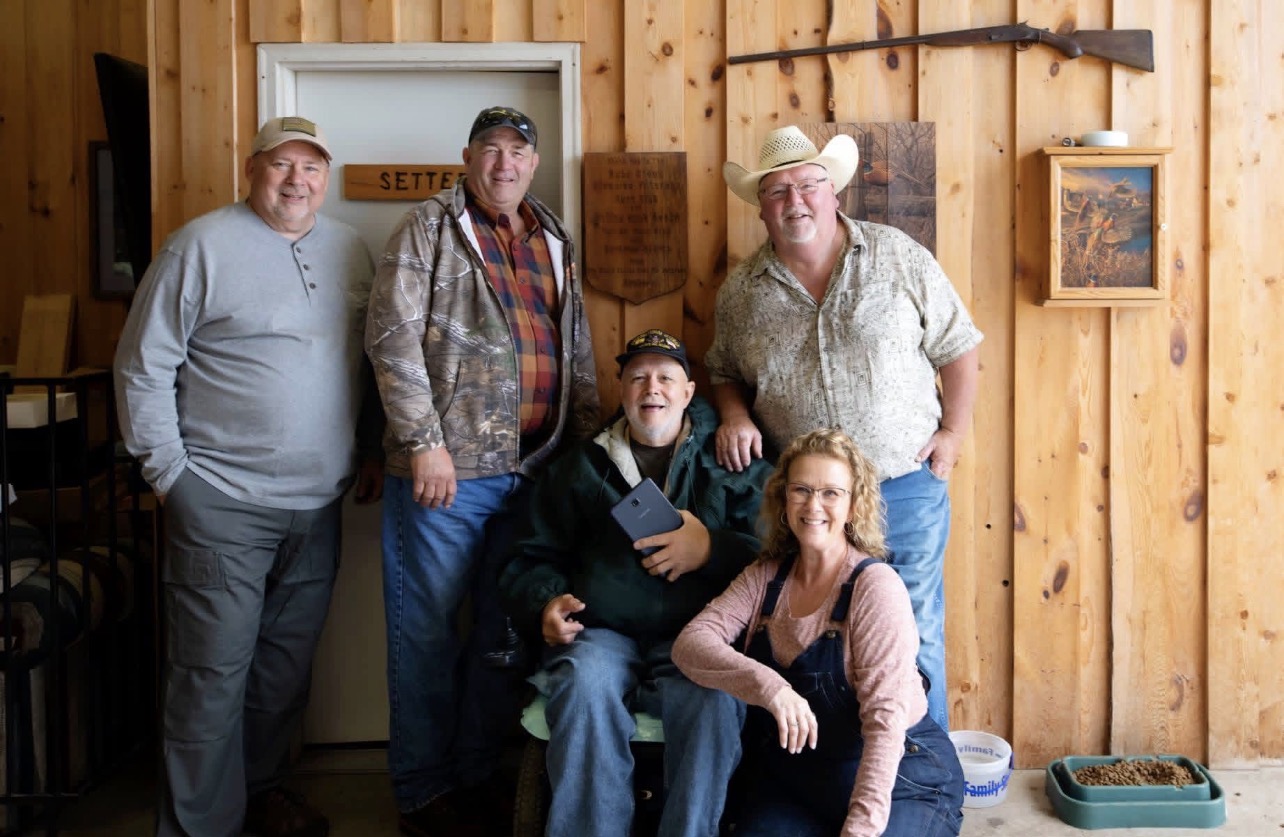 Five people, three standing and two sitting, pose inside a wooden room with framed pictures on the wall.