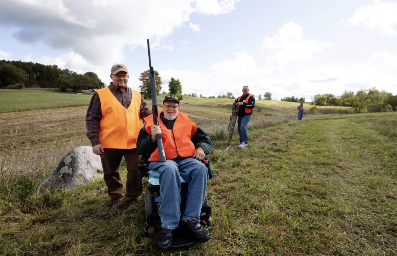 Three people outdoors in a grassy field, two wearing orange vests, one seated in a wheelchair holding a fishing rod.
