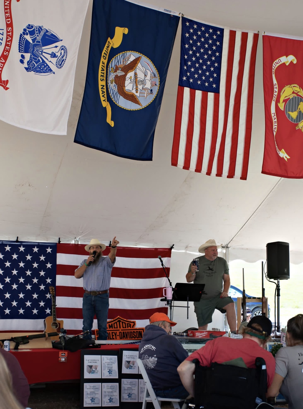 People on stage with American flags and military flags hanging above, audience seated in front, indoor event setting.