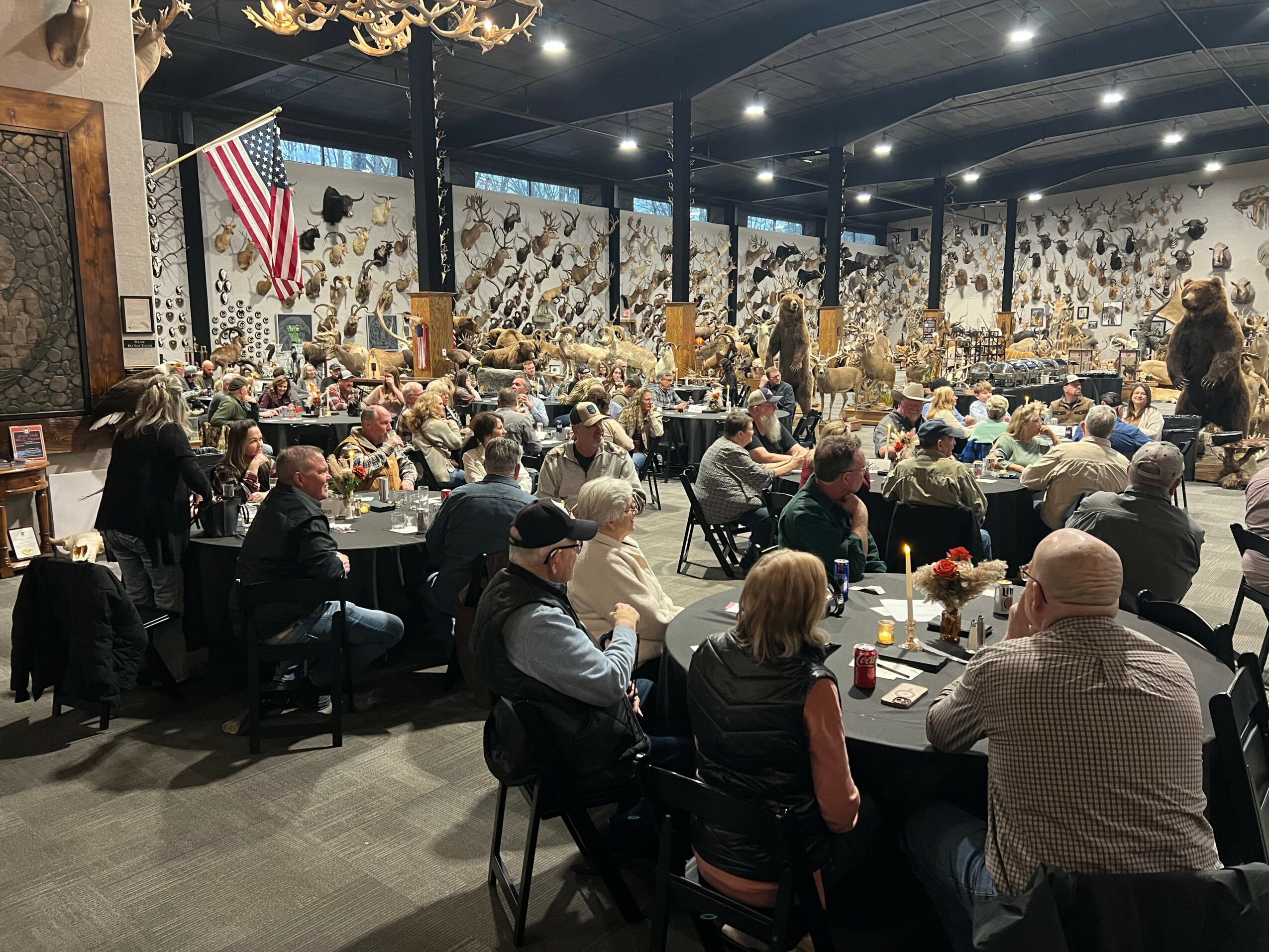 Large indoor gathering with many people seated at tables, some standing, in a decorated hall with an American flag.
