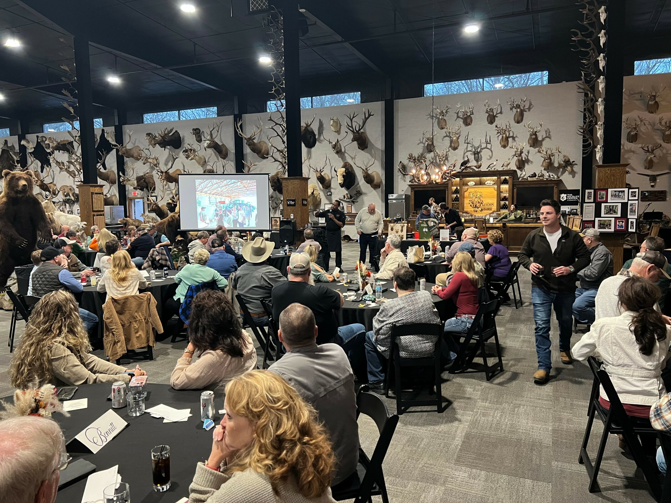 People seated at tables in a large room with display panels and a screen, some standing and talking, some seated and listening.