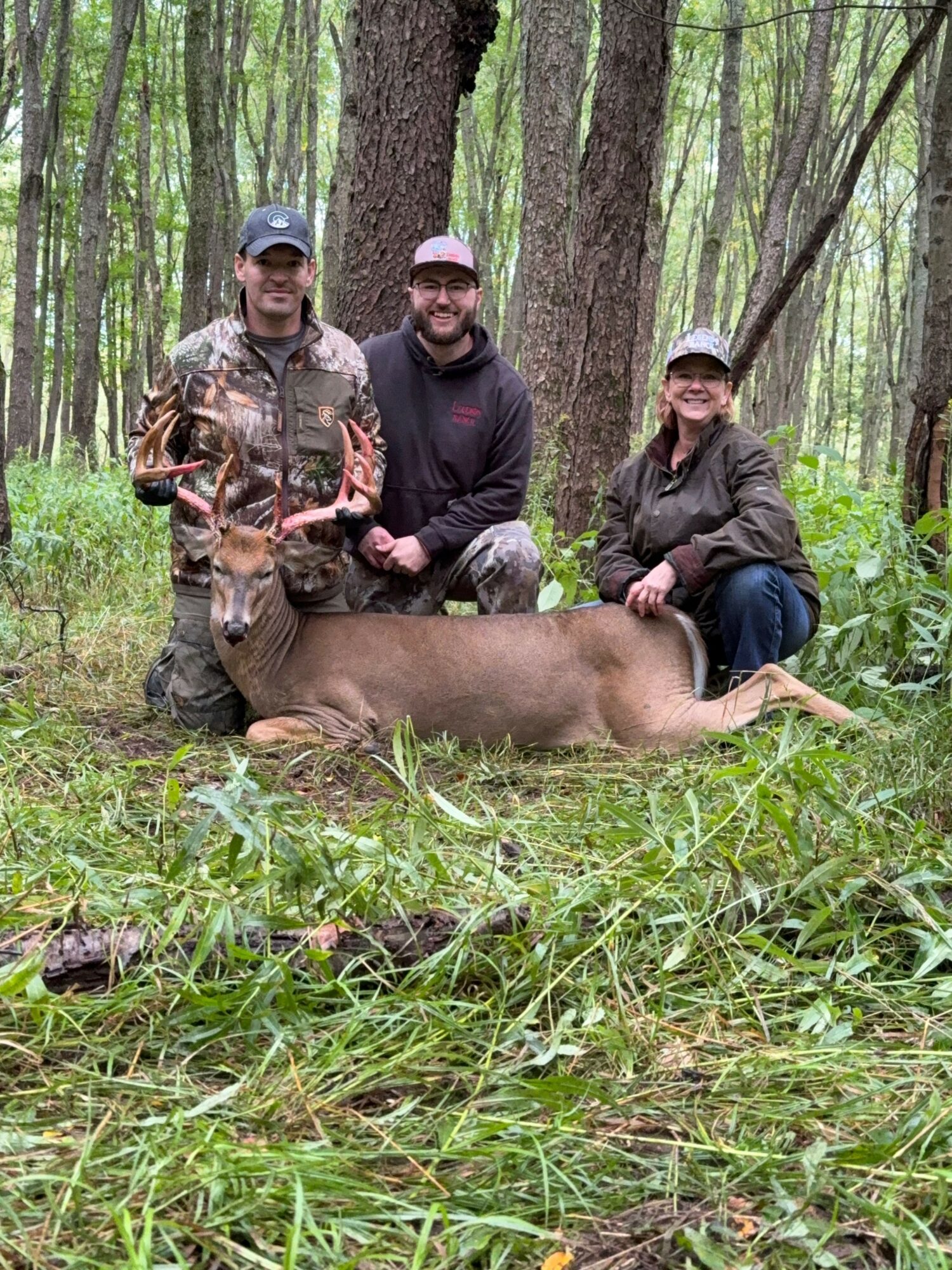 Three people kneeling behind a large, lying deer in a forest with green trees and grass.