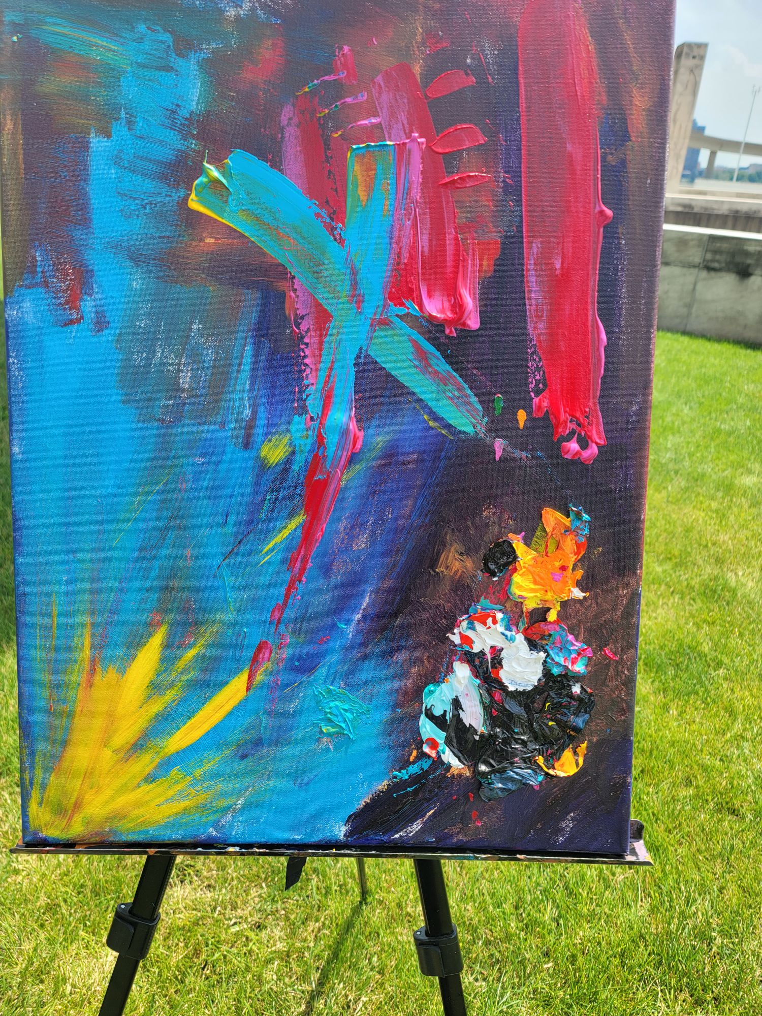 Abstract painting with blue, pink, yellow, and black colors on canvas, displayed outdoors on a black easel.