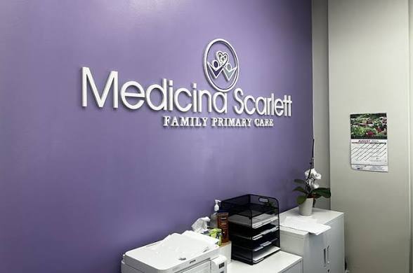 Reception area with purple wall, sign reading 'Medicina Scarlett Family Primary Care,' and a white counter with office supplies.