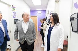 Group of people walking and talking in a brightly lit hallway, including a woman in a white coat and a man in a gray blazer.