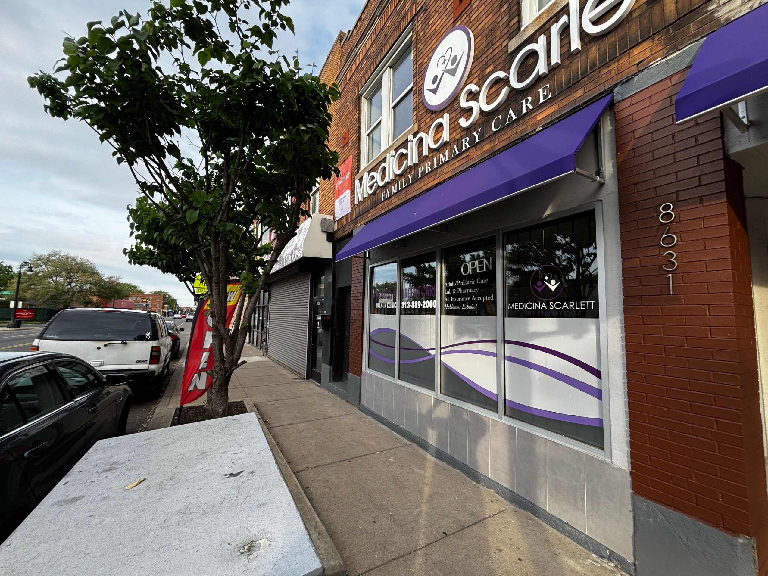 Street view of a storefront with purple awning, glass windows, and a sign reading 'Medicina Scarlett.' Sidewalk and parked cars are visible.