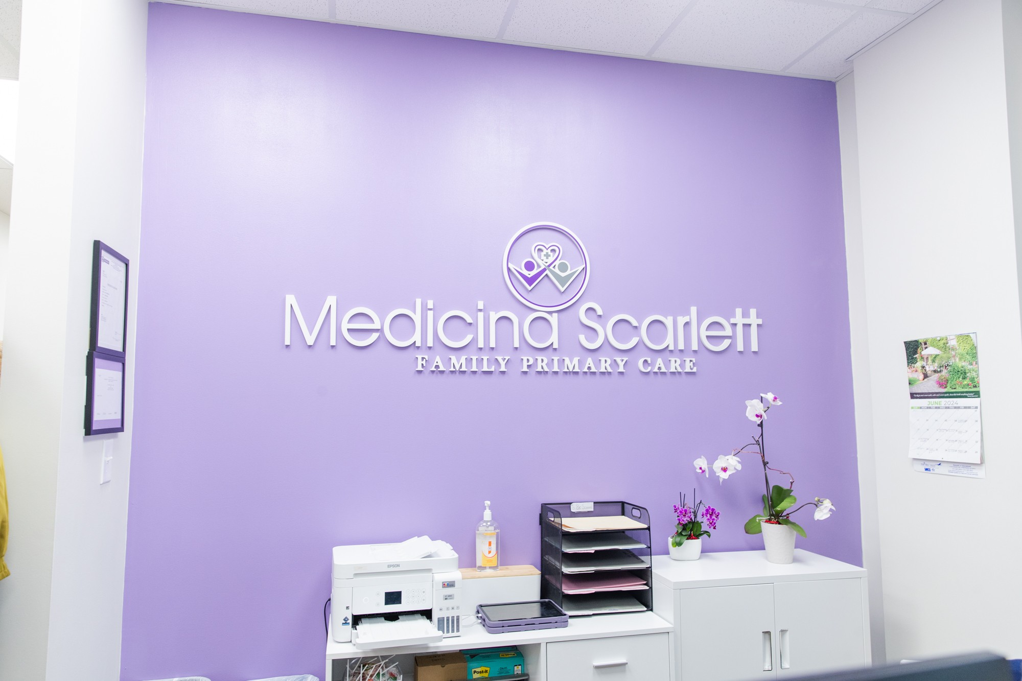 Reception area with purple wall, sign reading 'Medicine Scarlett Family Primary Care,' and white cabinet with flowers.