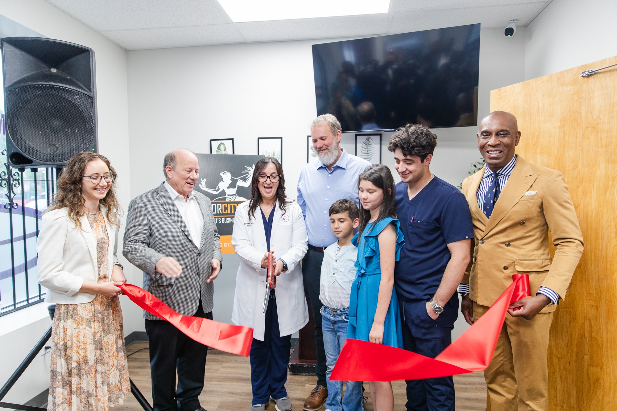 Group of nine diverse people at ribbon-cutting ceremony indoors, smiling, with a large screen and speaker in background.