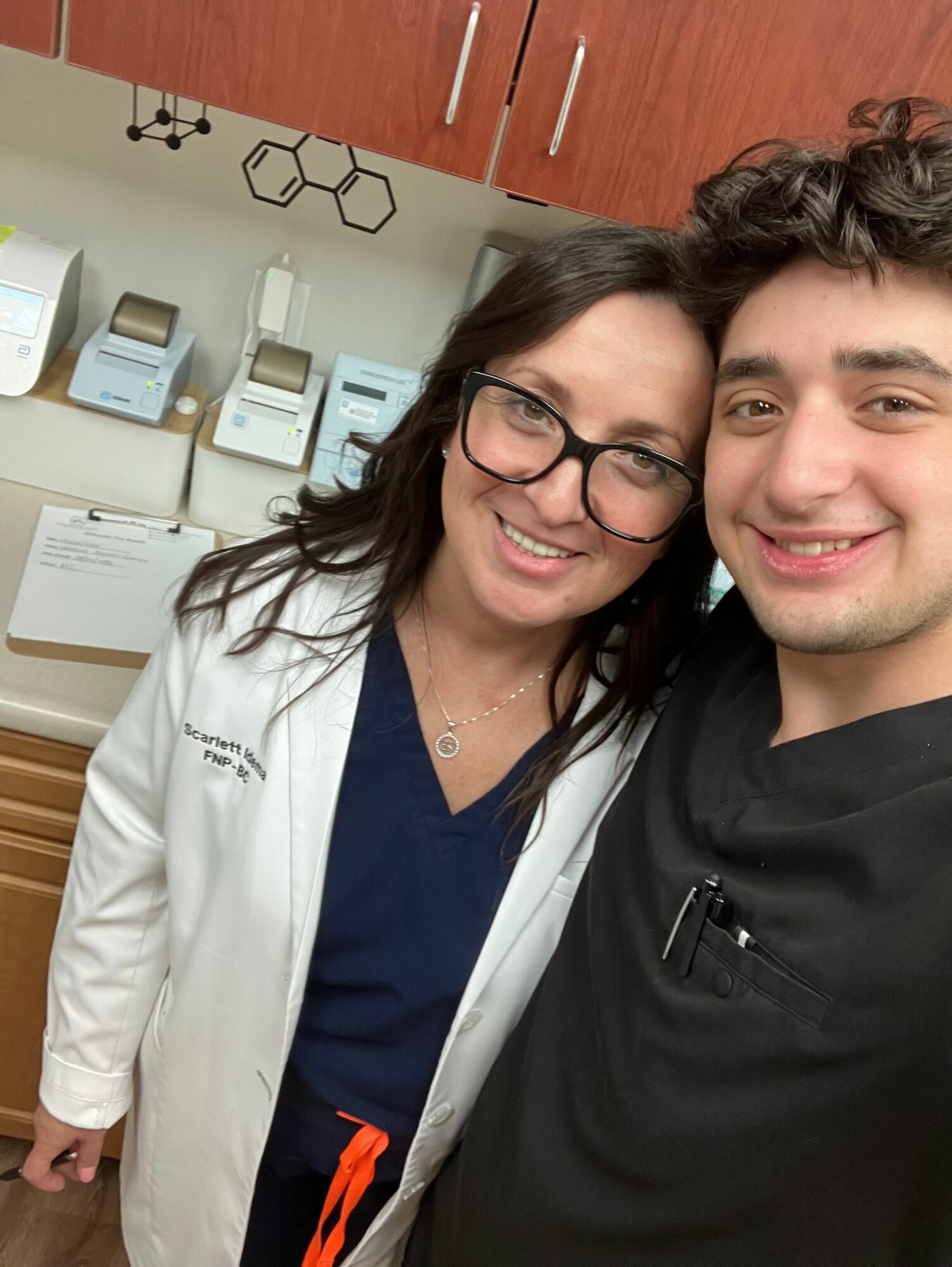 Two people smiling, one wearing glasses and a white coat, the other in a black outfit, in a medical setting.