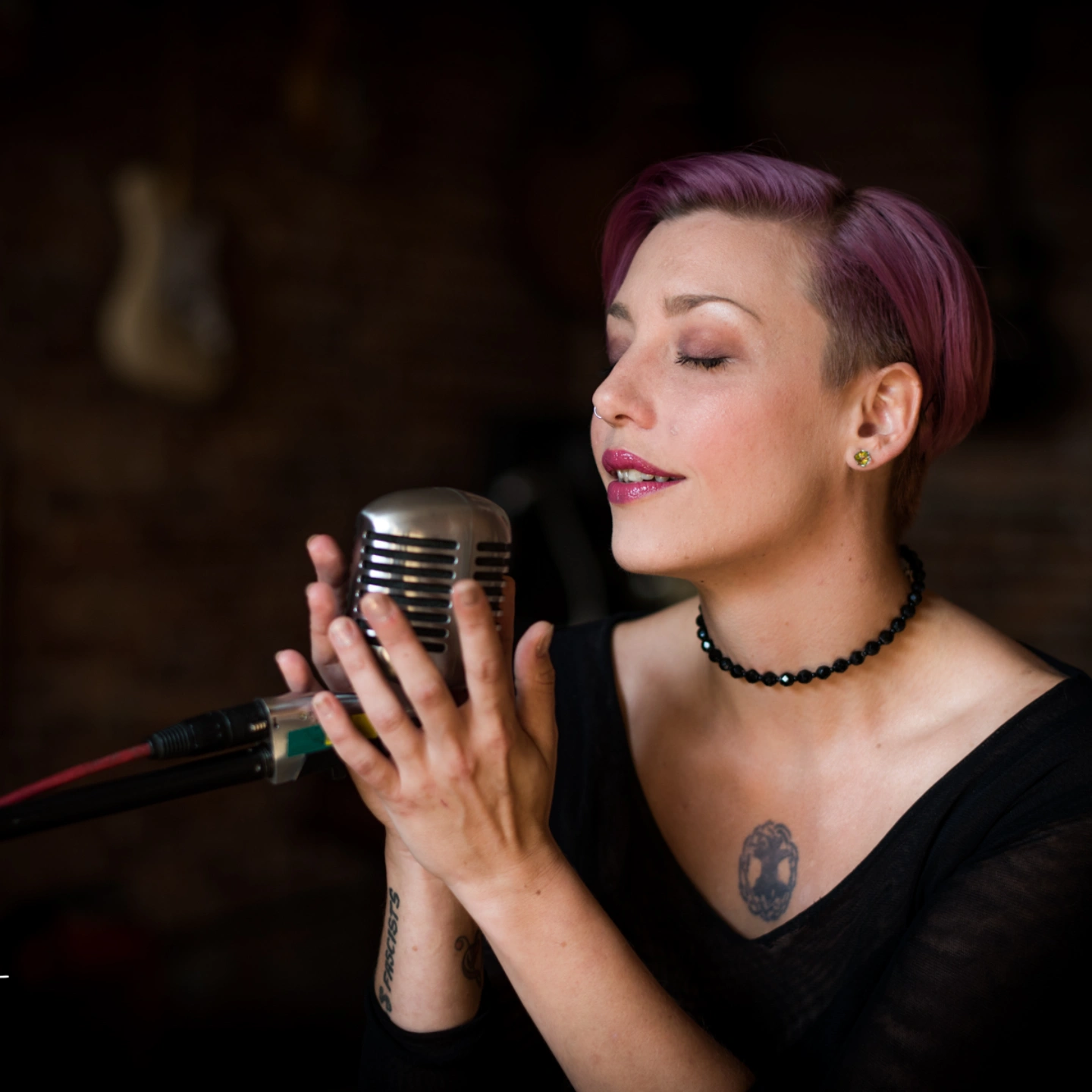 Woman with short purple hair singing into a vintage microphone, eyes closed, in a dimly lit setting.