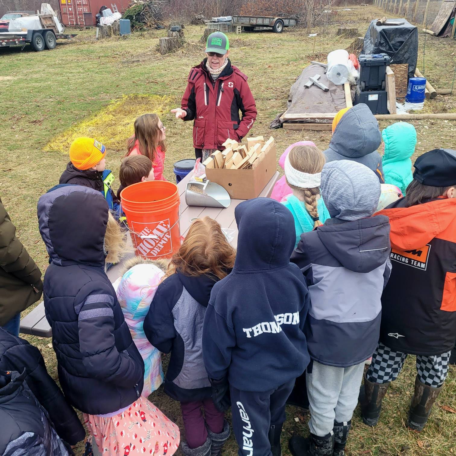 Group of children gathered outdoors around a woman in a red jacket, with trees and equipment in the background.
