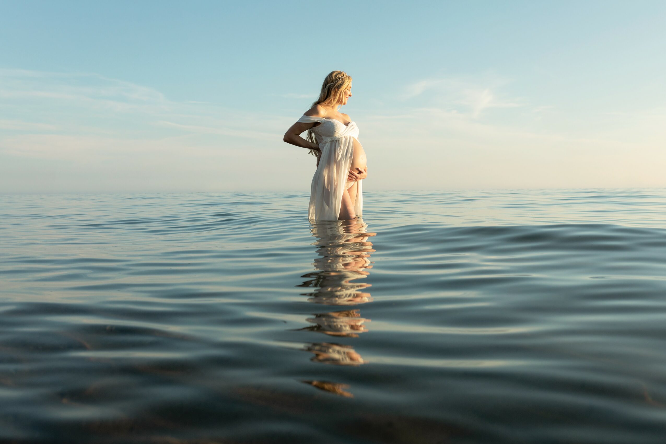 Woman standing in water with sky and horizon in background, wearing a light dress, looking to the side.