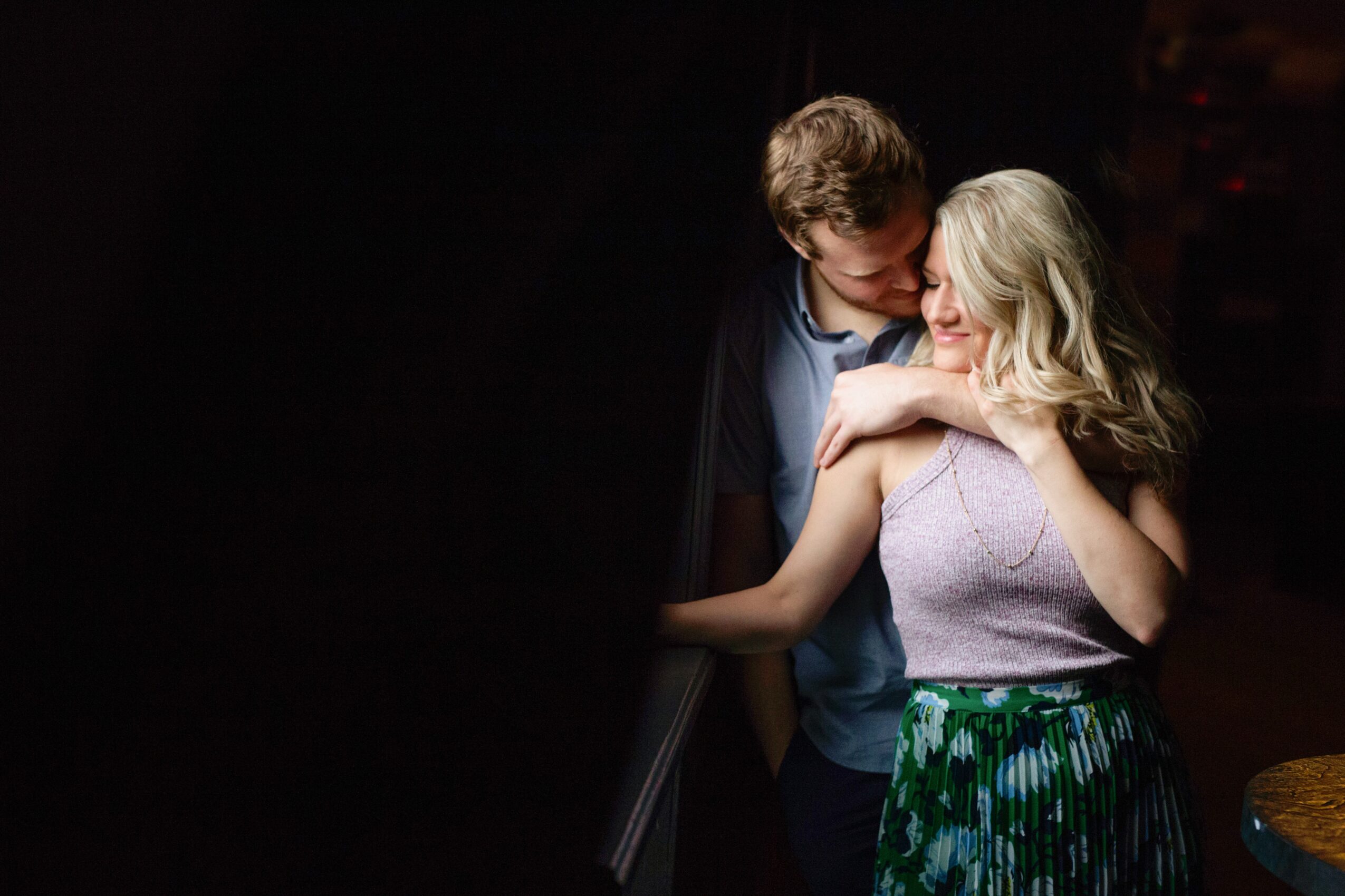 A young man and woman embrace in a dark setting, with the woman smiling and the man leaning close.