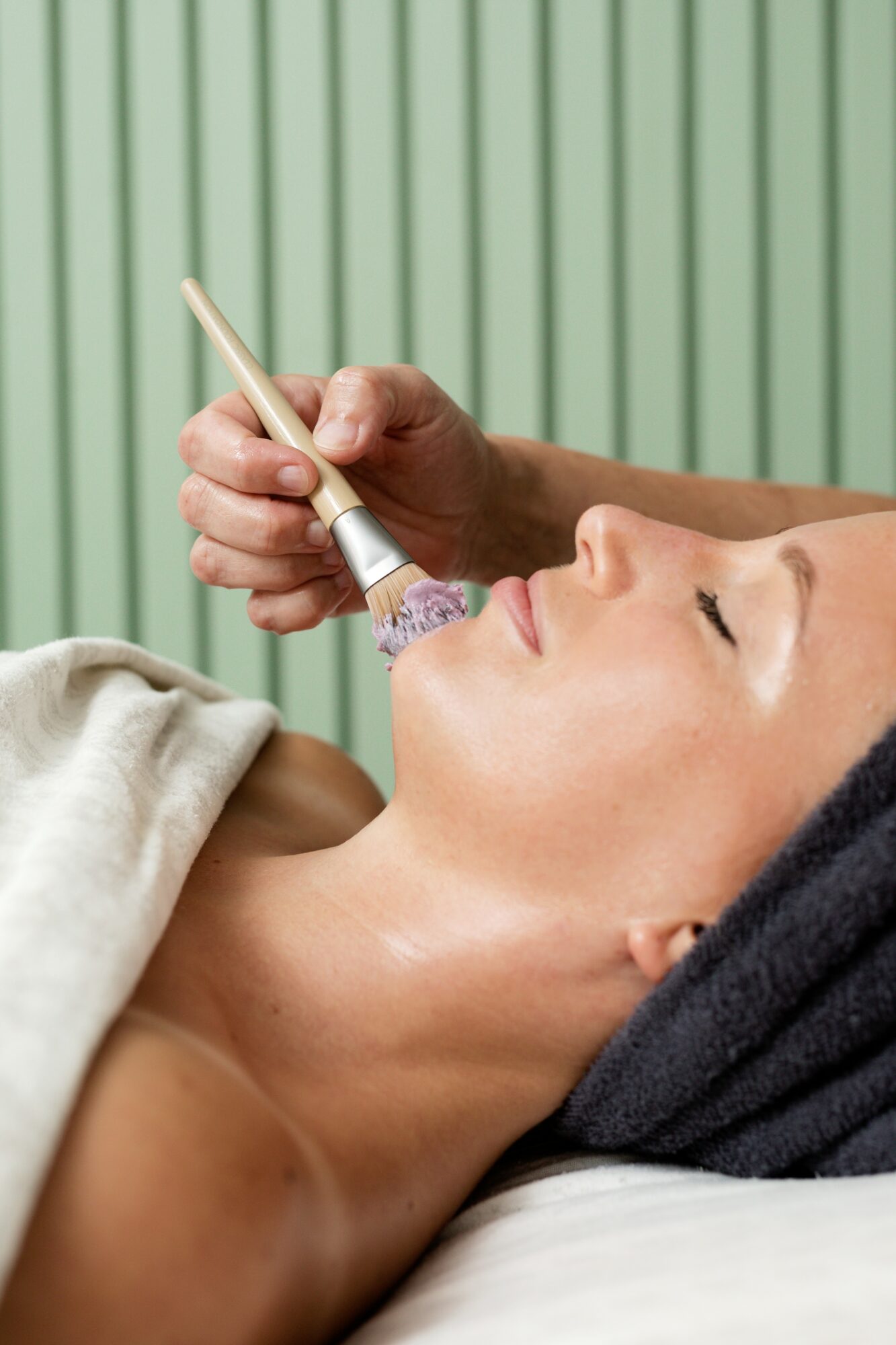 Woman lying down receiving facial treatment with a brush, green background with vertical lines.