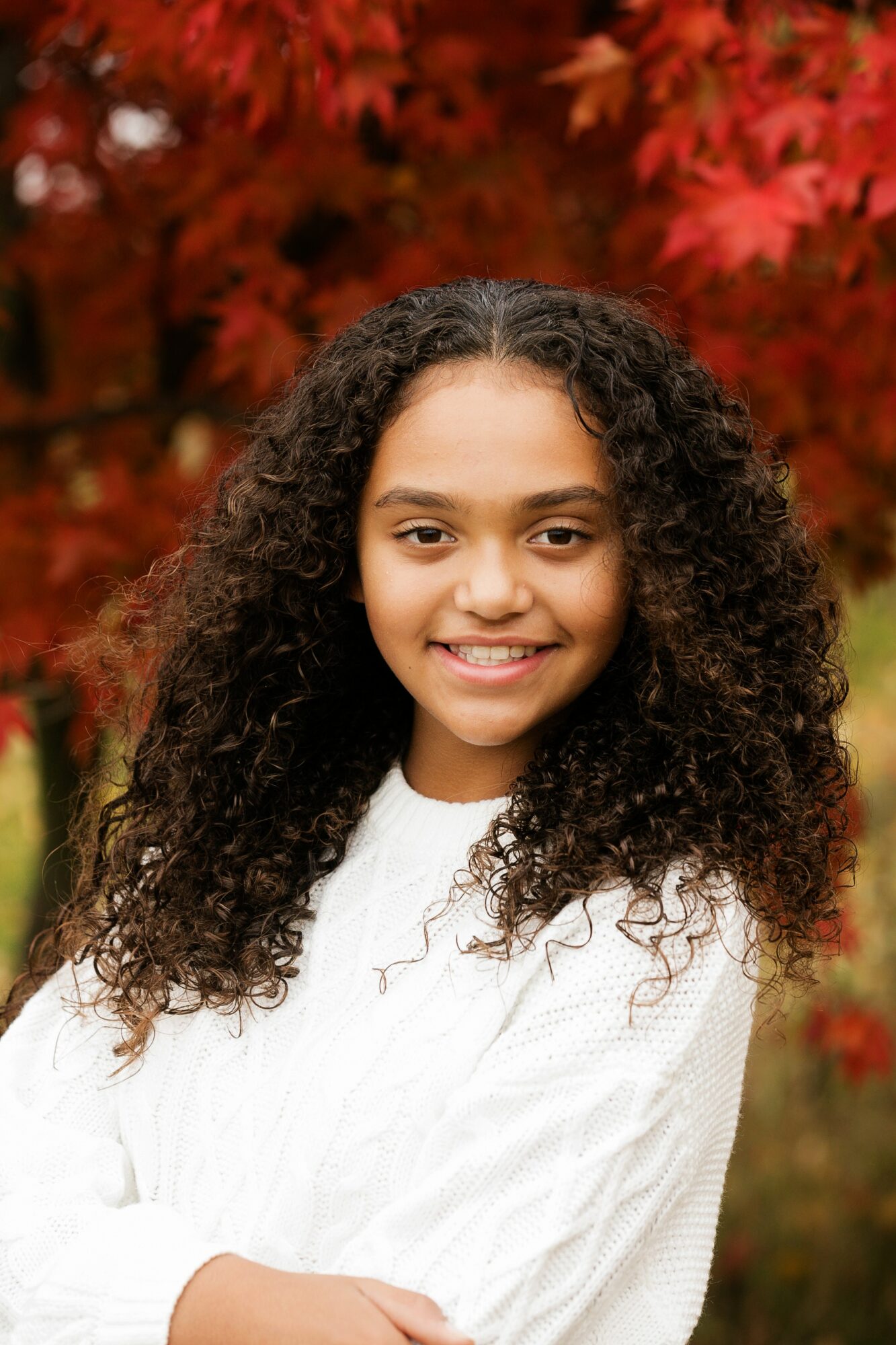 Young girl with curly hair smiling outdoors with red autumn leaves in background.