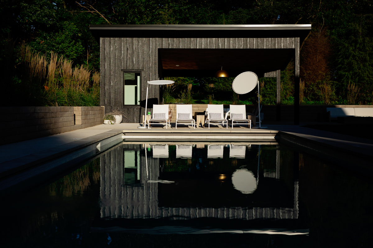 Modern outdoor pool area with lounge chairs, umbrellas, and a covered patio, reflecting in the water.