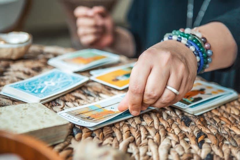 Person's hand with bracelets and ring, selecting tarot cards on woven surface, with other cards and a notebook nearby.