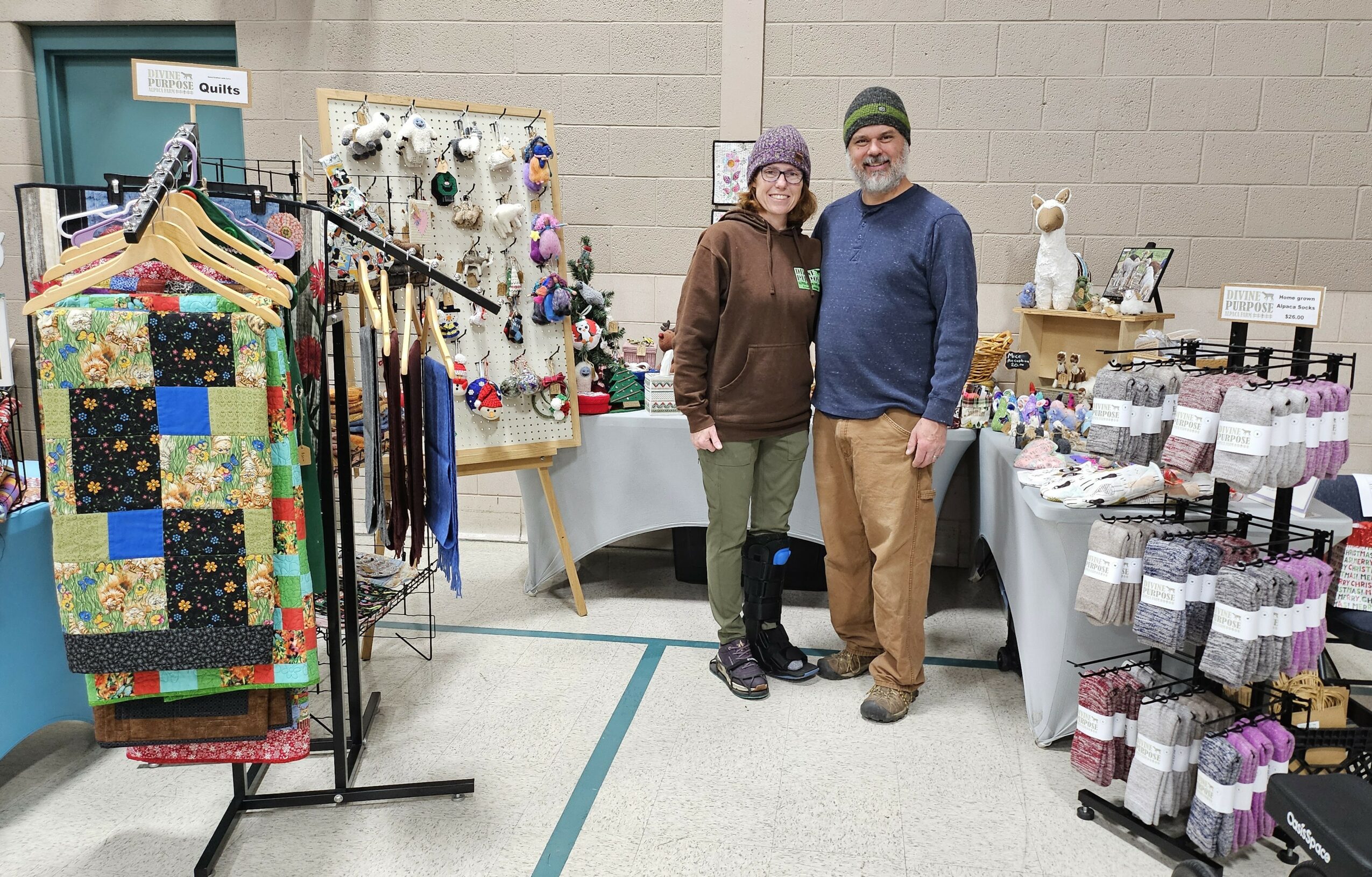 Two people stand in front of a display of handmade crafts and jewelry at an indoor market.
