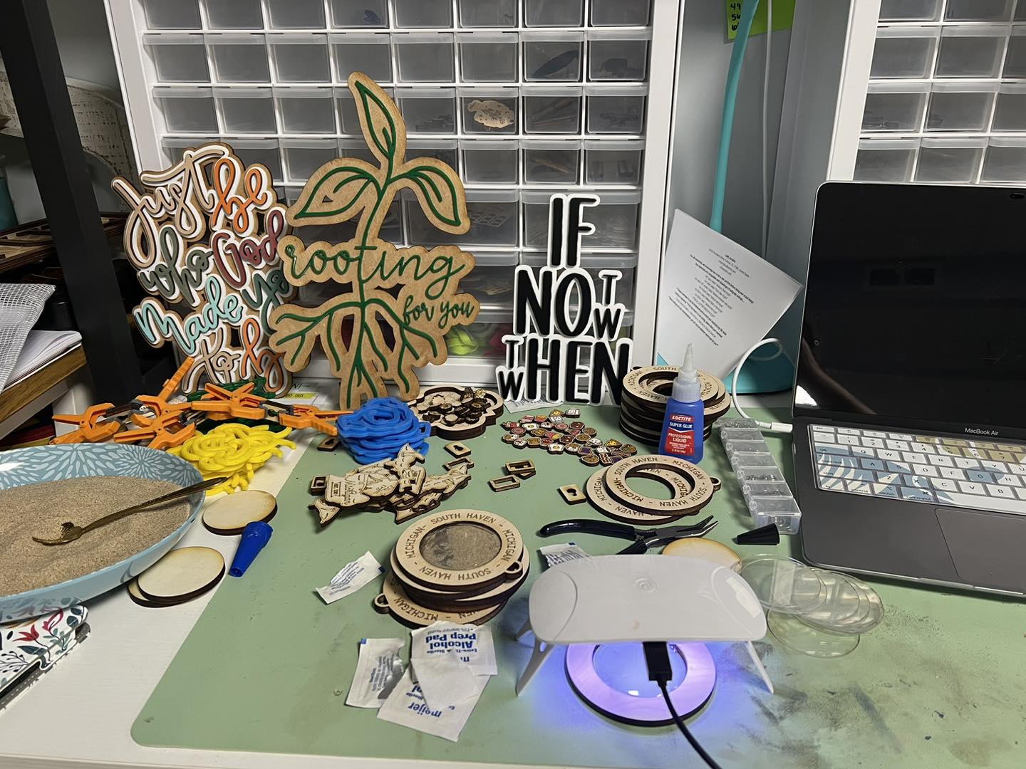 Workstation with craft supplies, a laptop, and a glowing device on a green mat, against a background of storage bins.