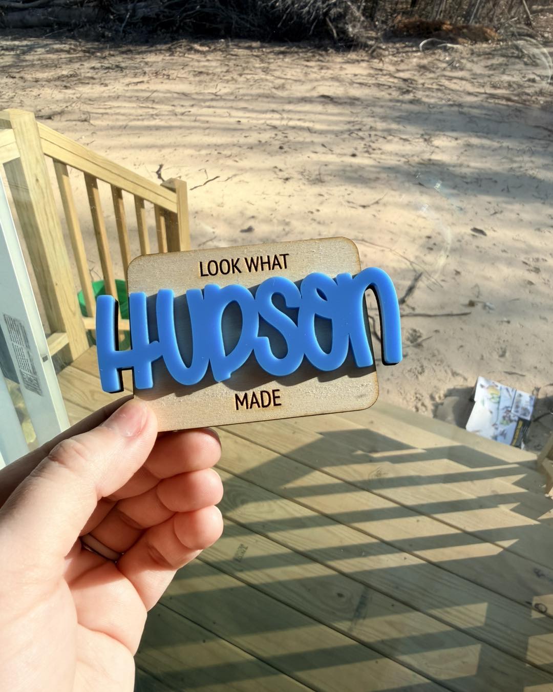 Hand holding a wooden sign with blue 3D letters spelling 'HUDSON' outdoors, sandy ground in background.
