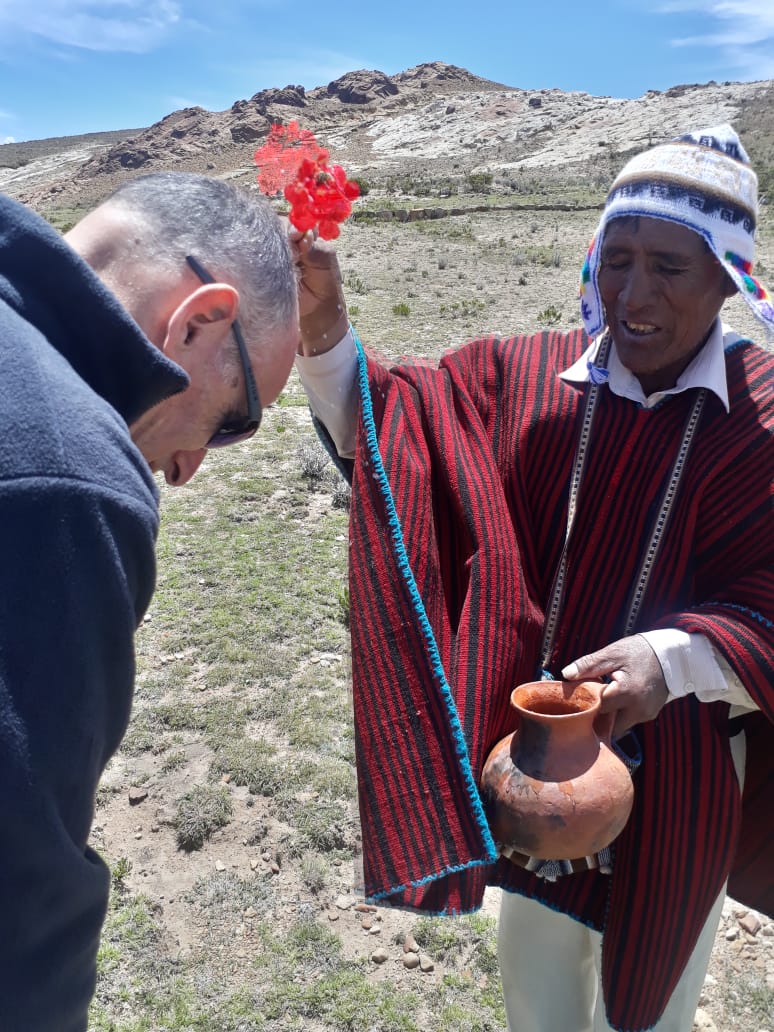 Person in traditional clothing holding a decorated string, another person bowing their head outdoors with mountains in background.