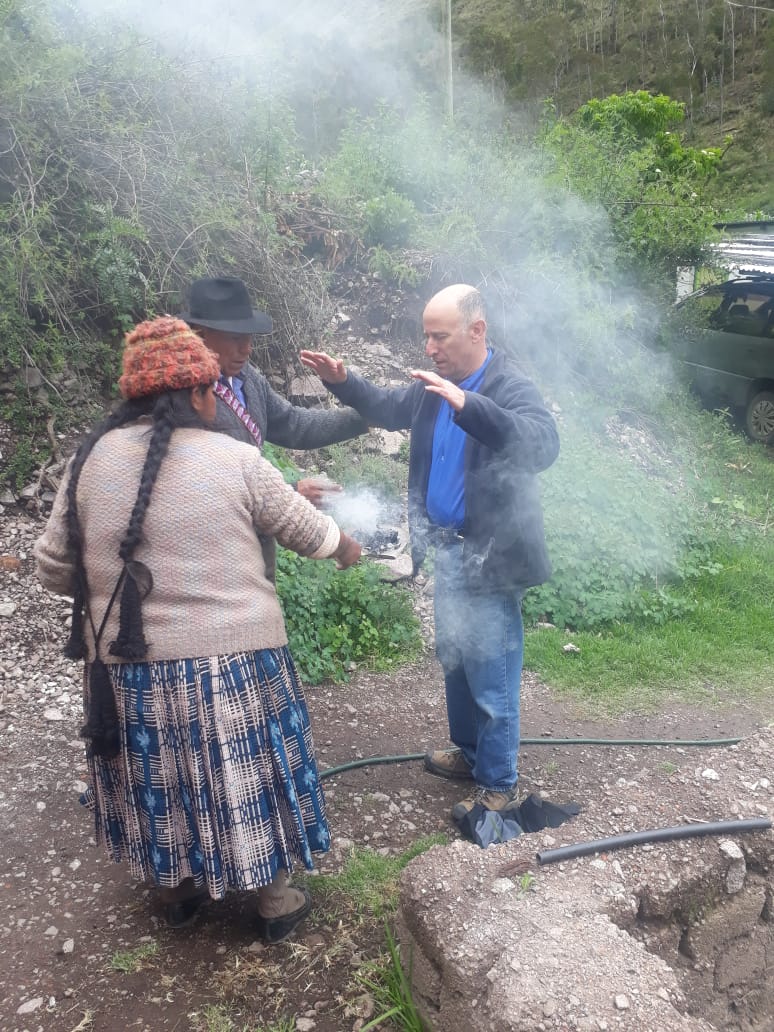 Four people standing outdoors near a smoky area, engaging in a ritual or activity, with greenery and a vehicle in the background.