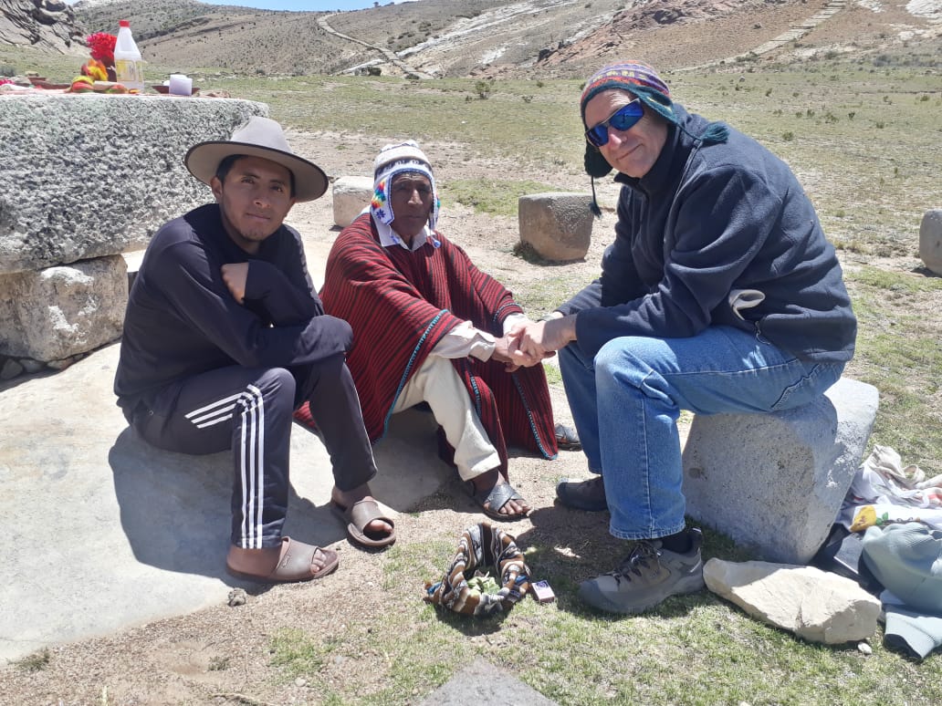 Three people sitting outdoors on rocks, two shaking hands, with a landscape of hills in the background.