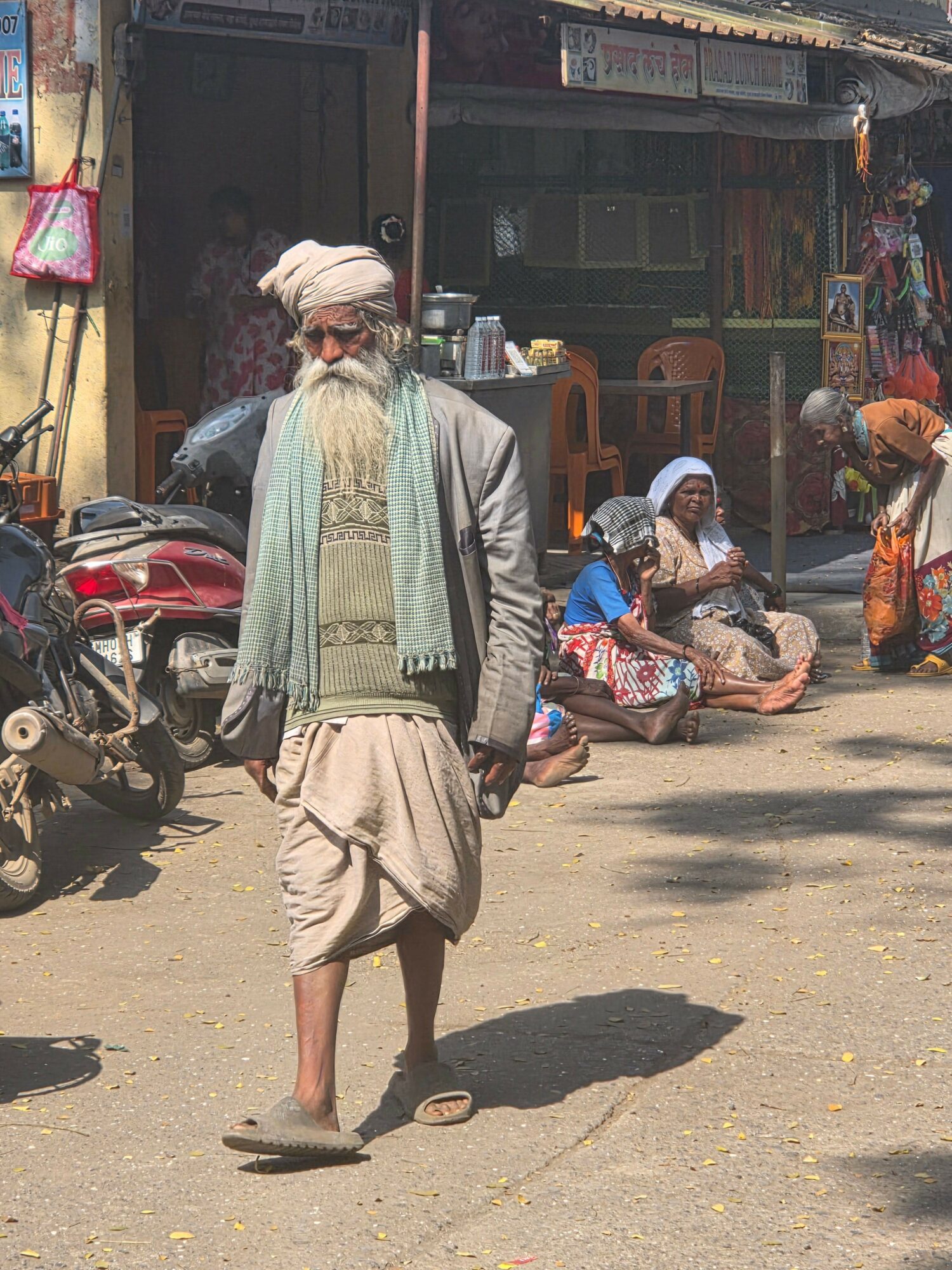 Elderly man with a long white beard and turban walking on street, people sitting and standing nearby, shops in background.