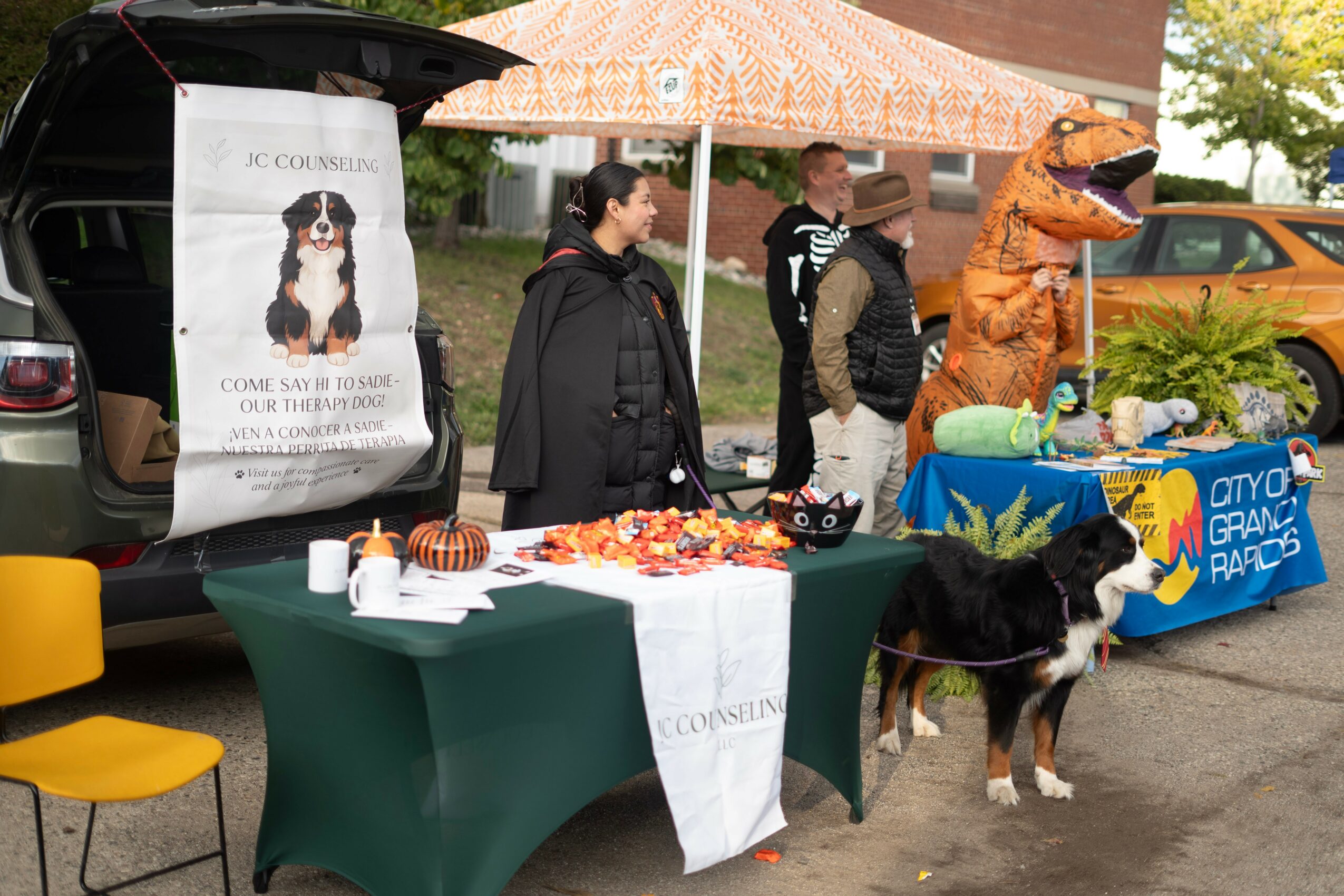 People stand near tables with promotional items, a person in a T-Rex costume, and a dog on a leash outdoors.