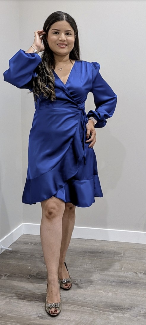Woman in blue dress standing against a plain wall, smiling, with one hand near her face and the other on her hip.