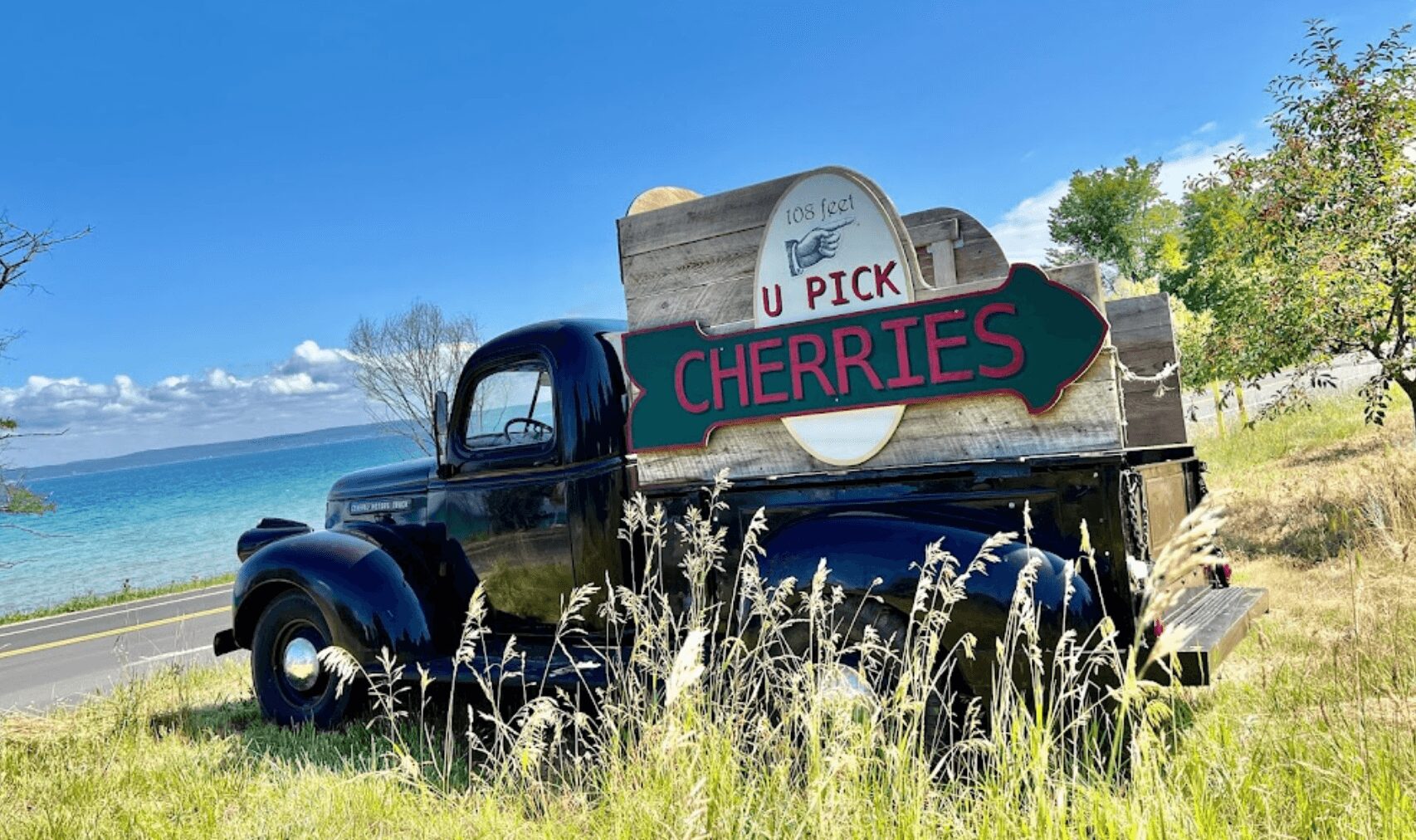 Old truck with a sign advertising U Pick cherries, surrounded by grass and trees under a blue sky.