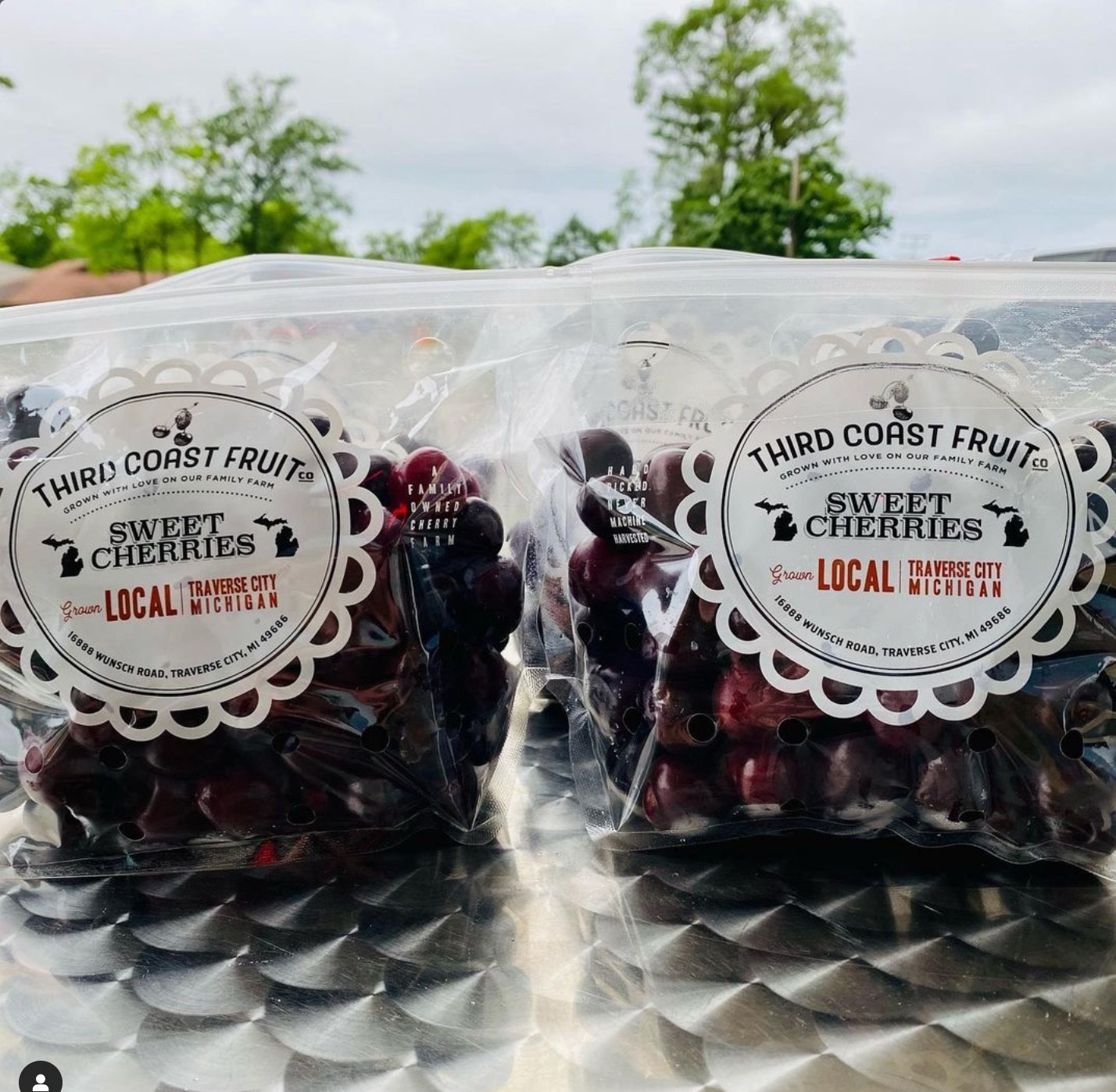 Two clear plastic bags filled with cherries, labels read 'Third Coast Fruit' and 'Sweet Cherries,' outdoors with trees and sky.
