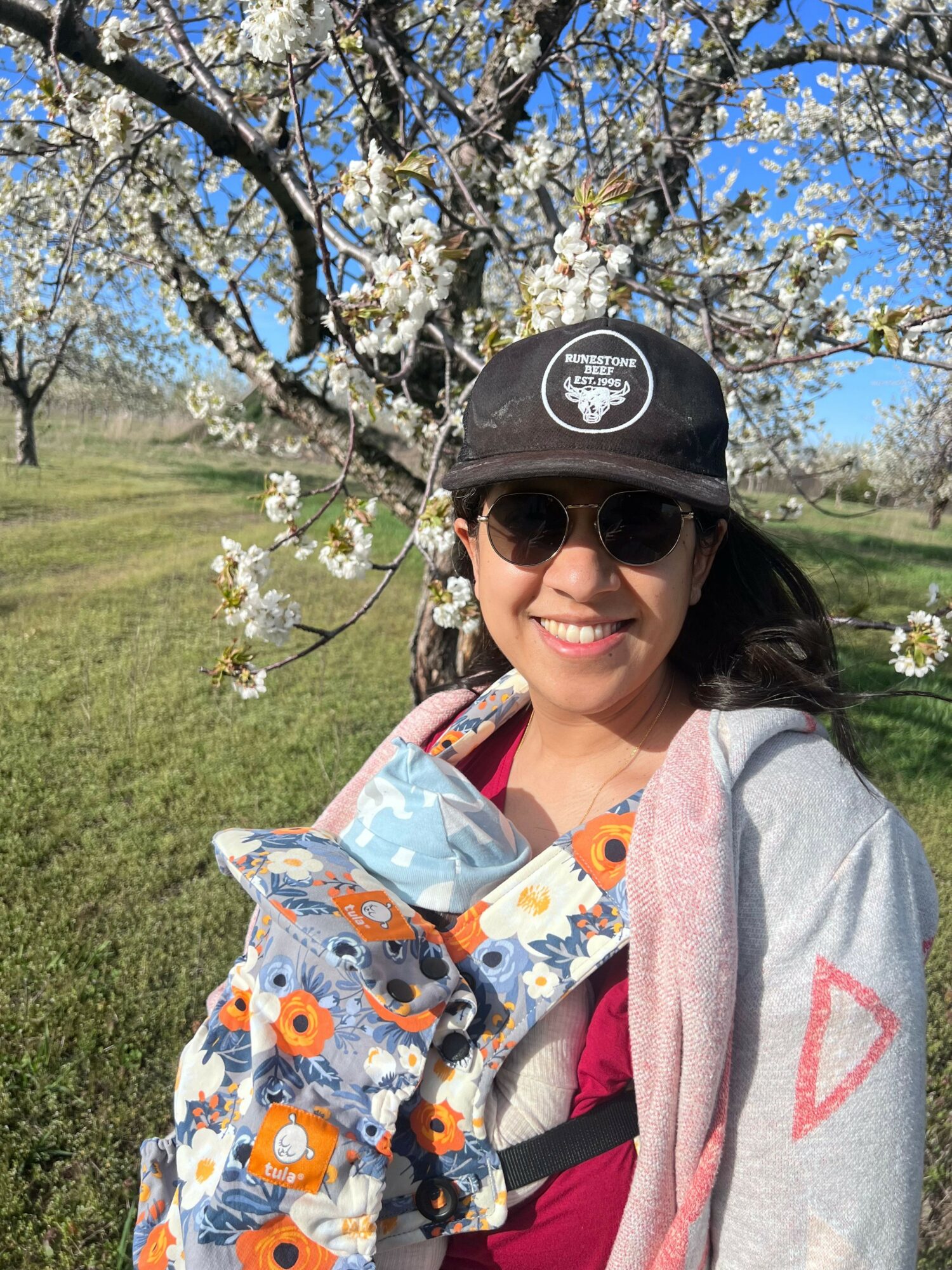 Woman smiling outdoors with sunglasses, black cap, floral backpack, pink scarf, and blooming tree behind her.