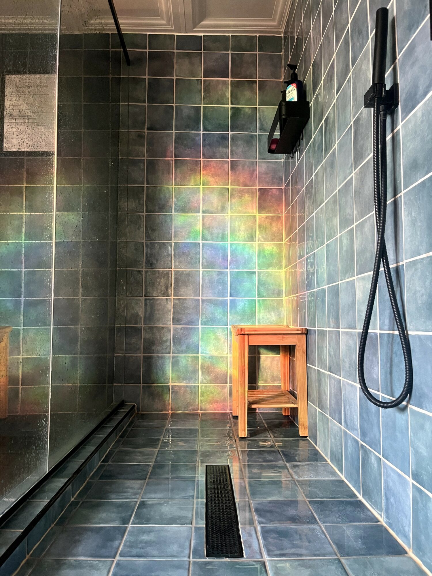 Shower area with tiled walls, a wooden stool, a black showerhead, and a rainbow reflection on the tiles.