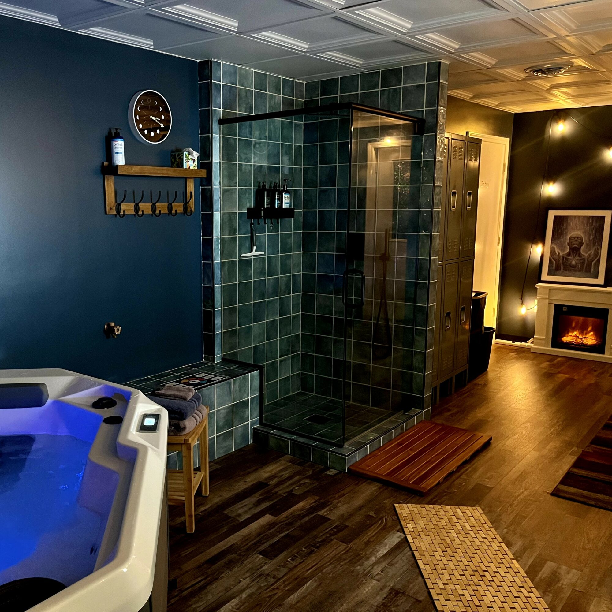 Bathroom with glass shower, blue wall, wooden shelf, clock, and a corner of a hot tub, warm lighting, wooden floor.