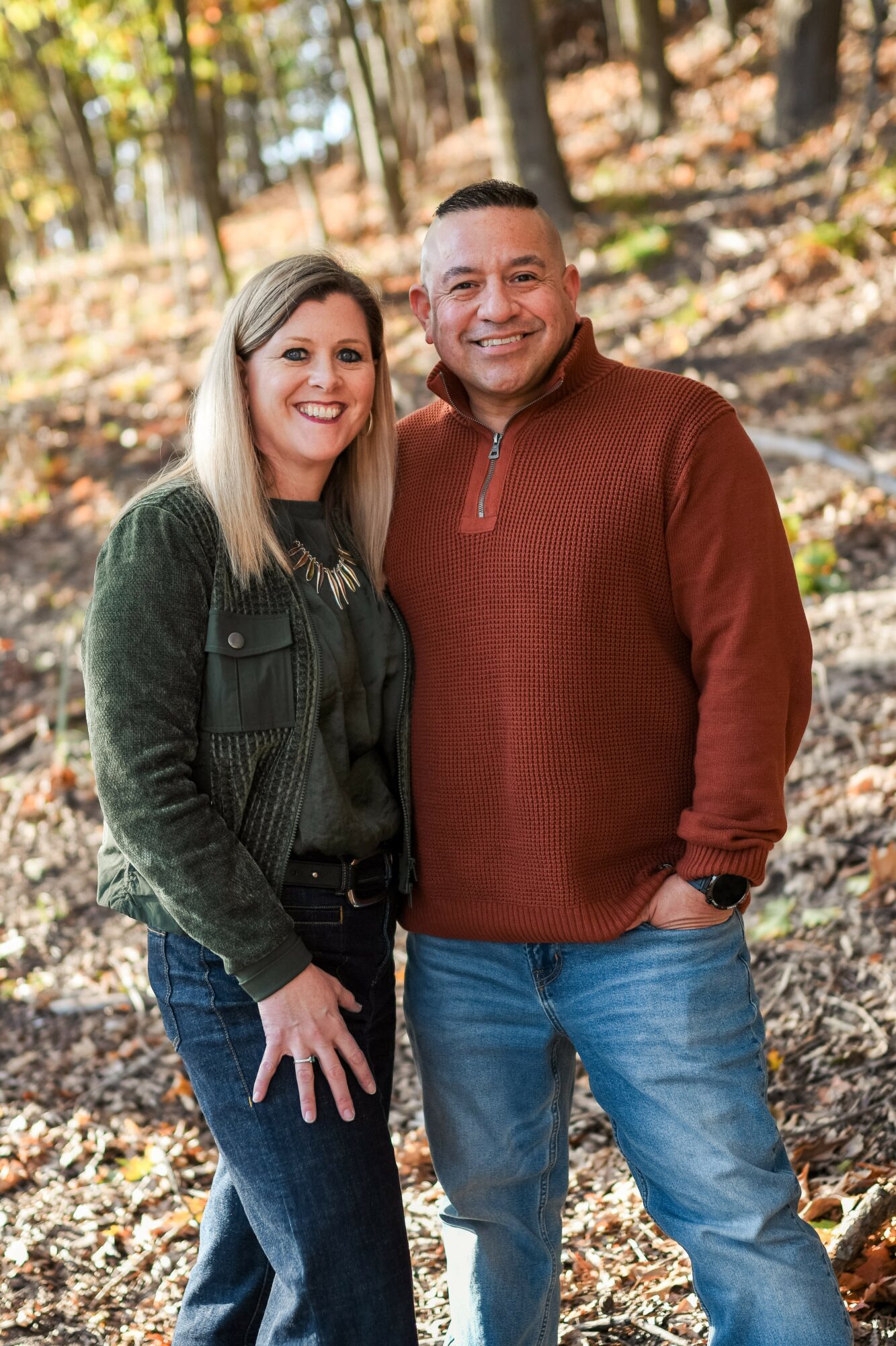 A smiling couple stands outdoors in a wooded area with fallen leaves, posing for a photo.