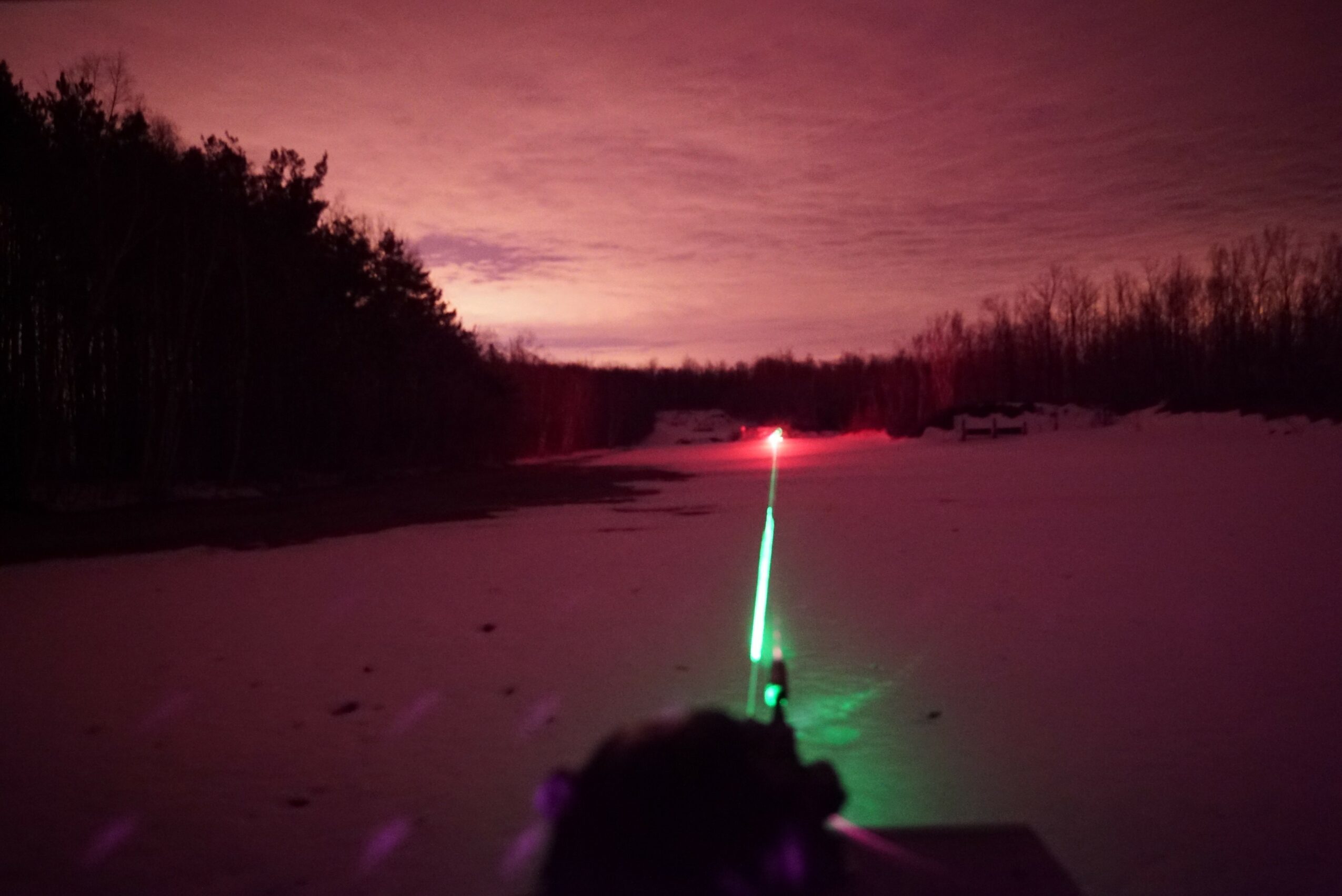 Snow-covered landscape with a glowing green and red light in the foreground, trees and a cloudy sky in the background.