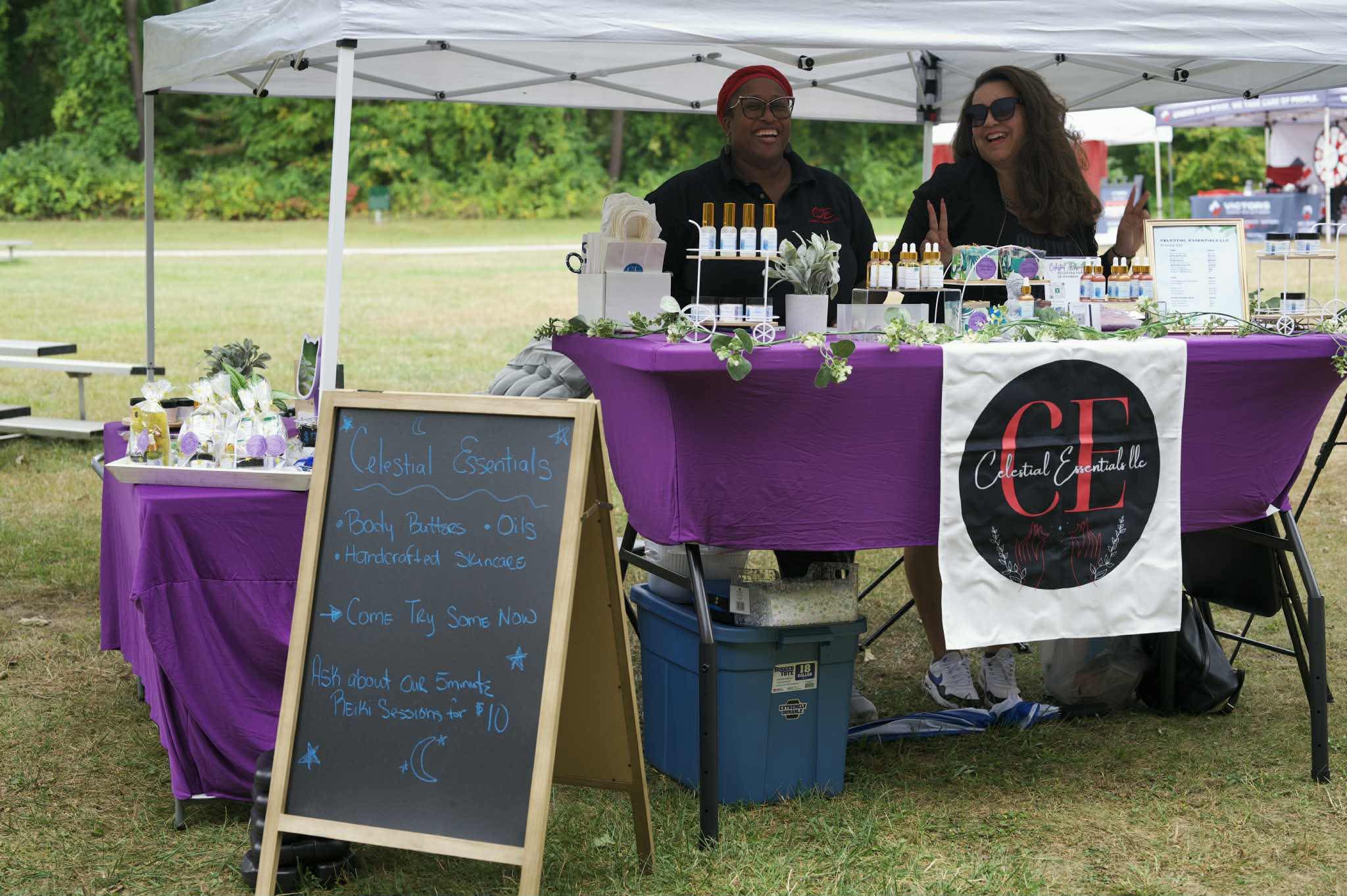 Two people stand behind a booth with a purple tablecloth under a white canopy, selling products outdoors.