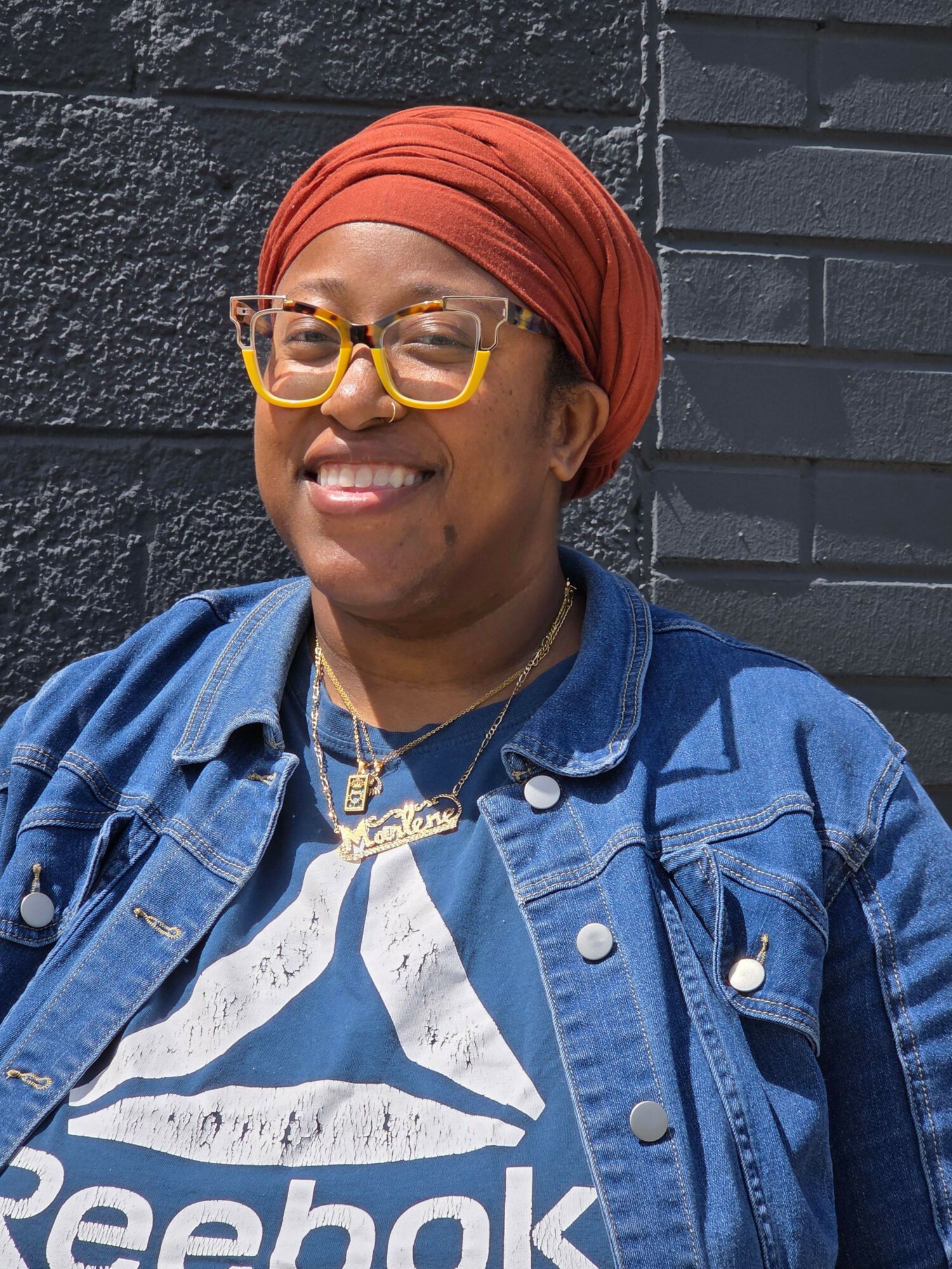 Woman wearing glasses, red headscarf, denim jacket, and Reebok shirt, smiling outdoors against a brick wall.