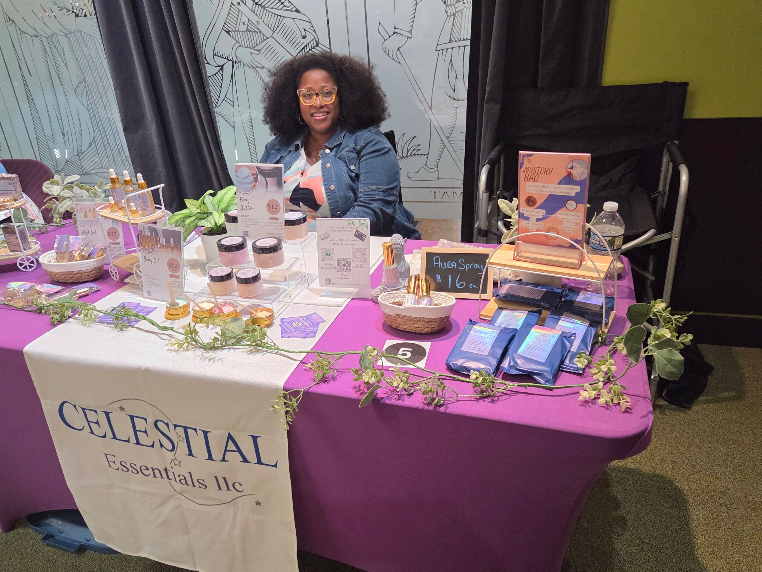 Woman sitting behind a table with skincare products and promotional materials, smiling, in an indoor setting.