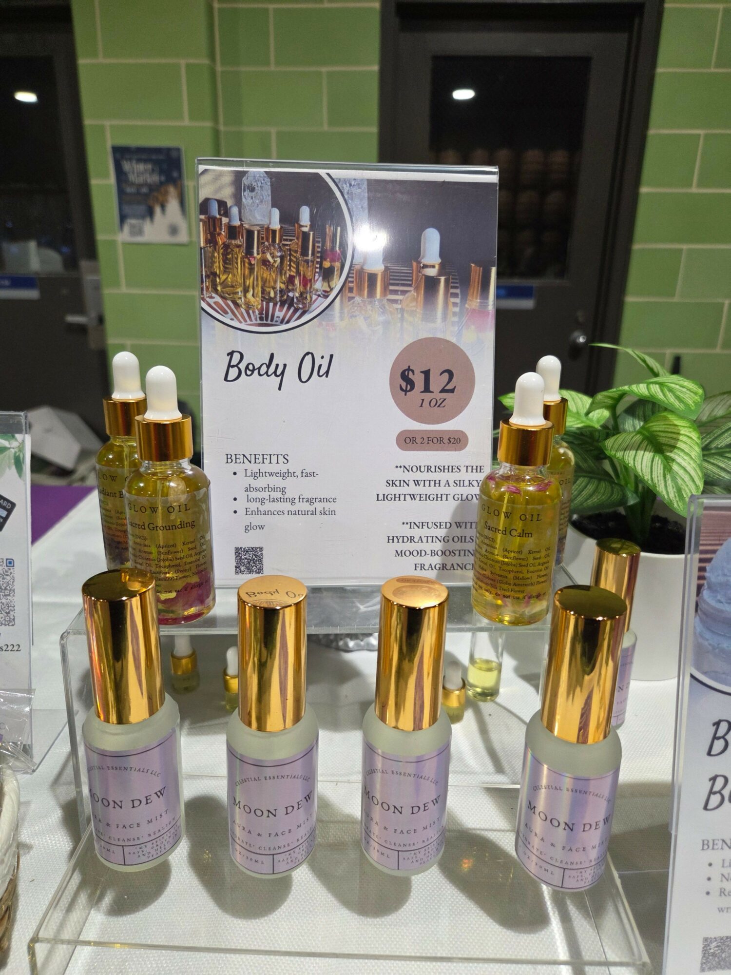Display of body oil bottles with gold caps and a sign in the background, green tiled wall, and a plant.