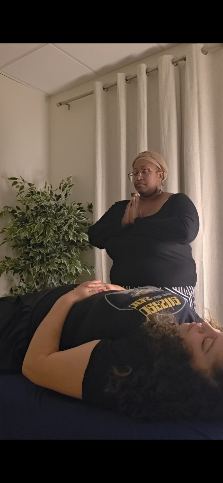 Person lying down with head turned to the side, another person standing nearby, in a room with curtains and a plant.