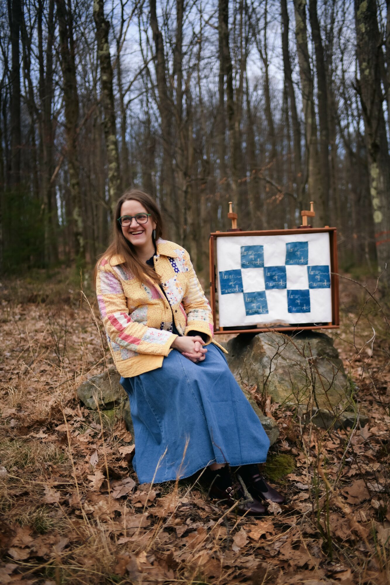 Woman sitting outdoors in a forest, smiling, with a chessboard on a stand beside her.