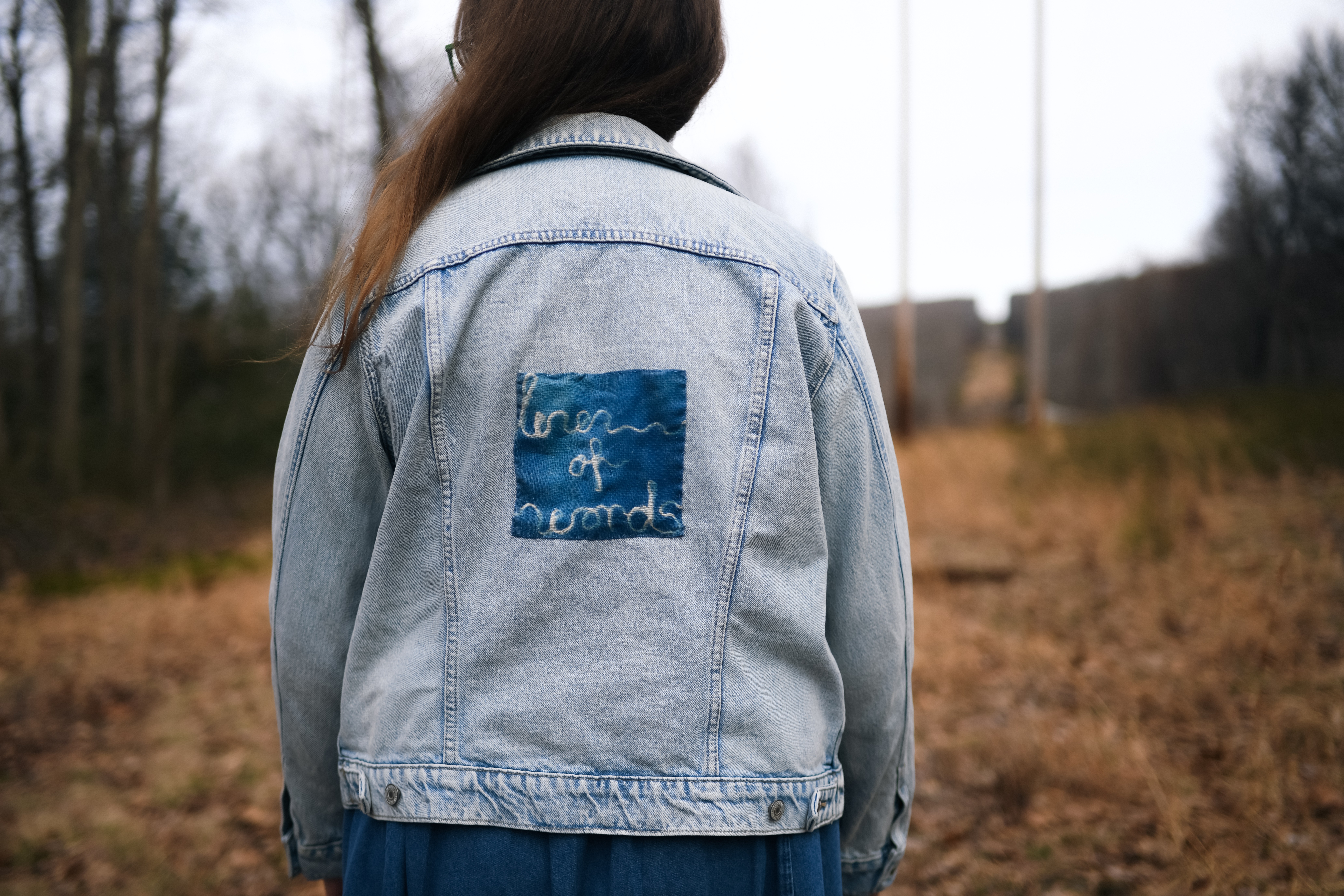 Person wearing a light blue denim jacket with a patch on the back, outdoors in a field.