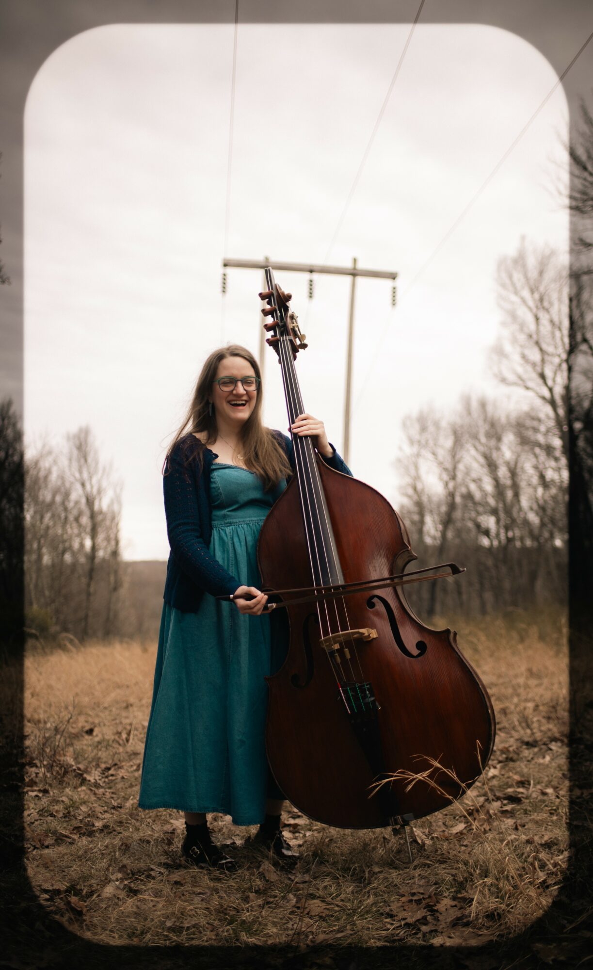 Woman standing outdoors holding a large double bass, with trees and power lines in the background.
